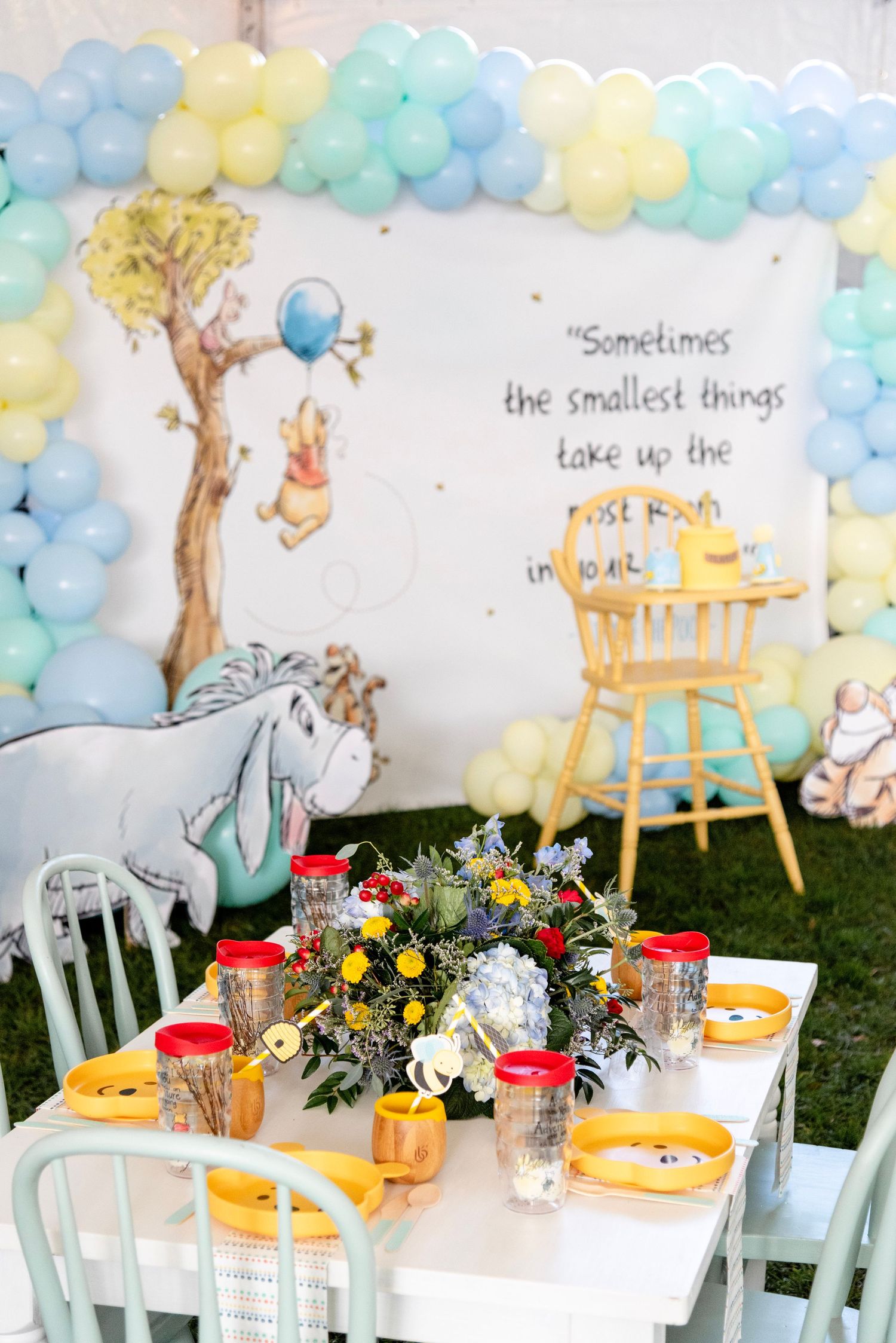 A whimsical baby shower table setting with pastel balloon garland, white chairs, and classic Winnie the Pooh decorations.