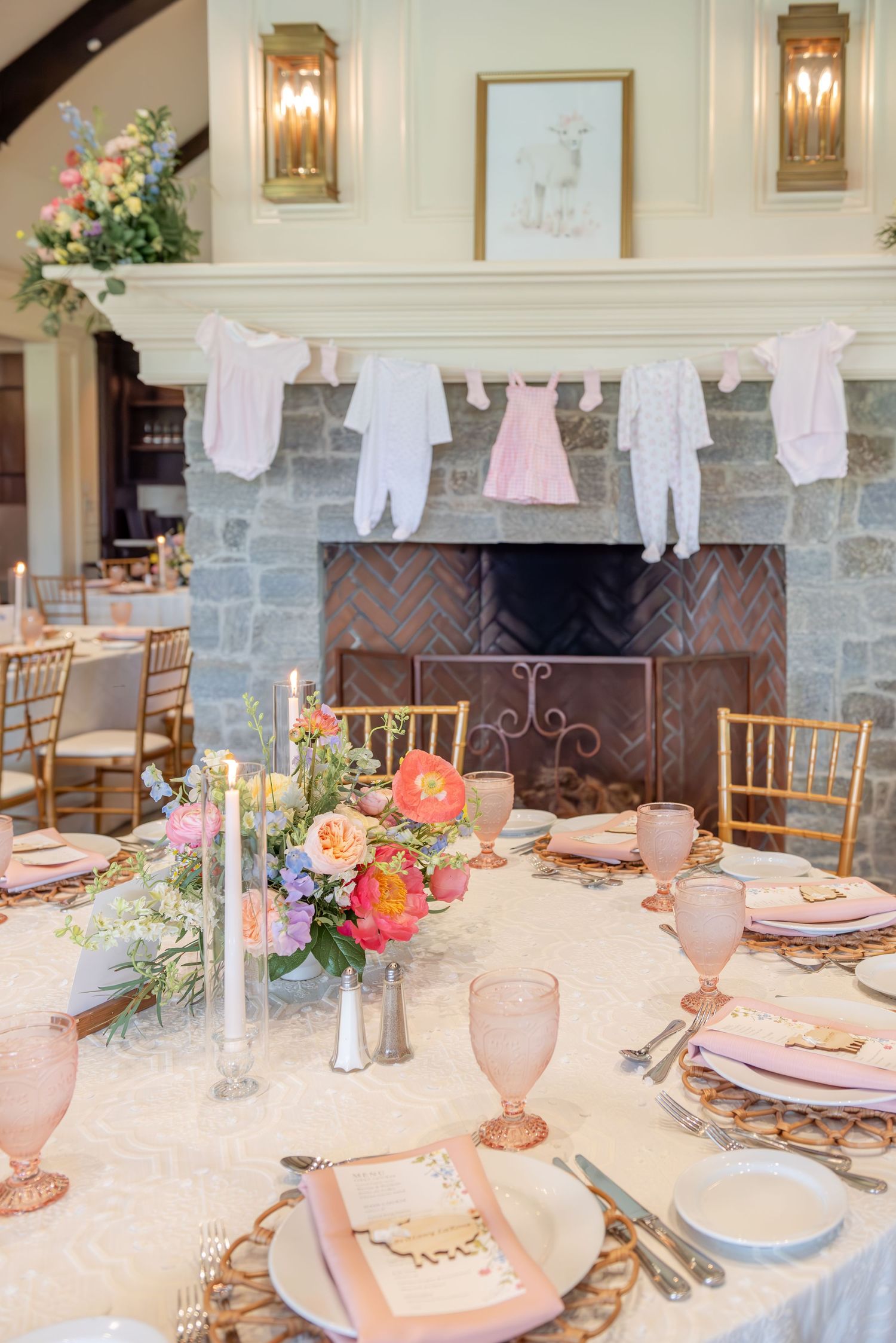 Elegant baby shower table settings with pink glassware, floral centerpieces, and baby clothes hanging as decor.