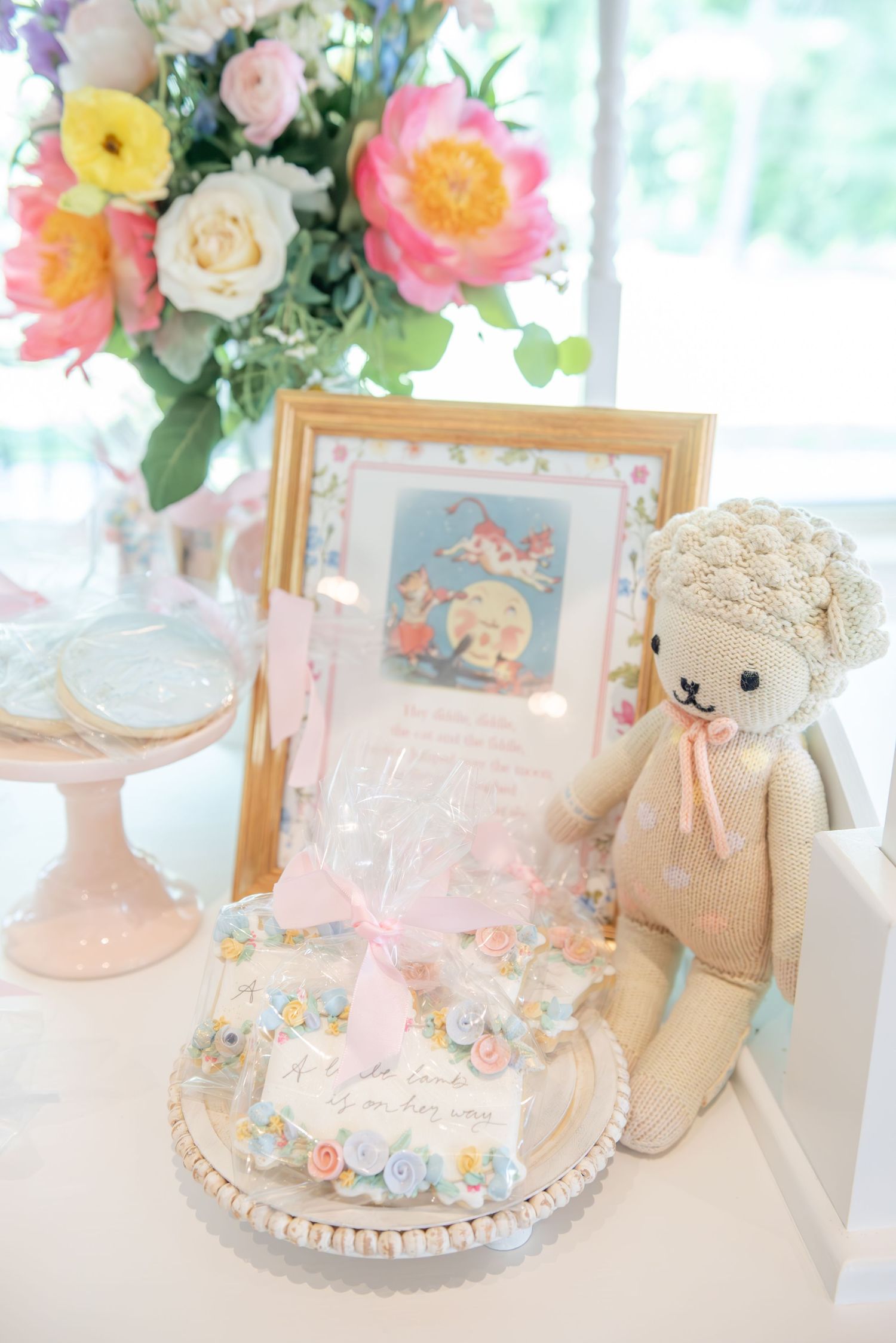 Elegant pastel party decor with floral arrangements, teddy bear and delicate cake displays on a white table.