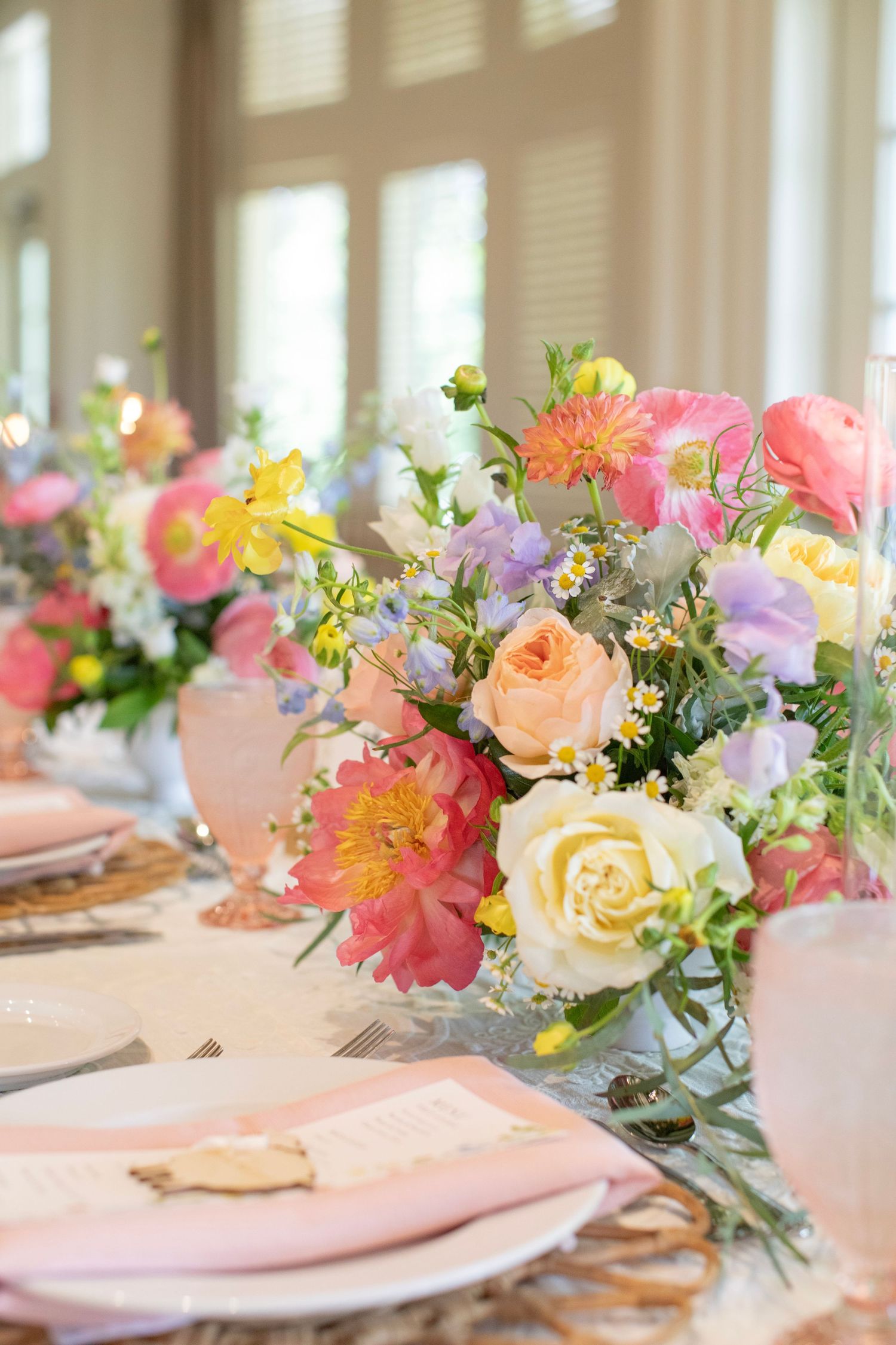 Elegant table setting with colorful floral centerpieces featuring pink, yellow and coral blooms on a soft pink tablecloth.