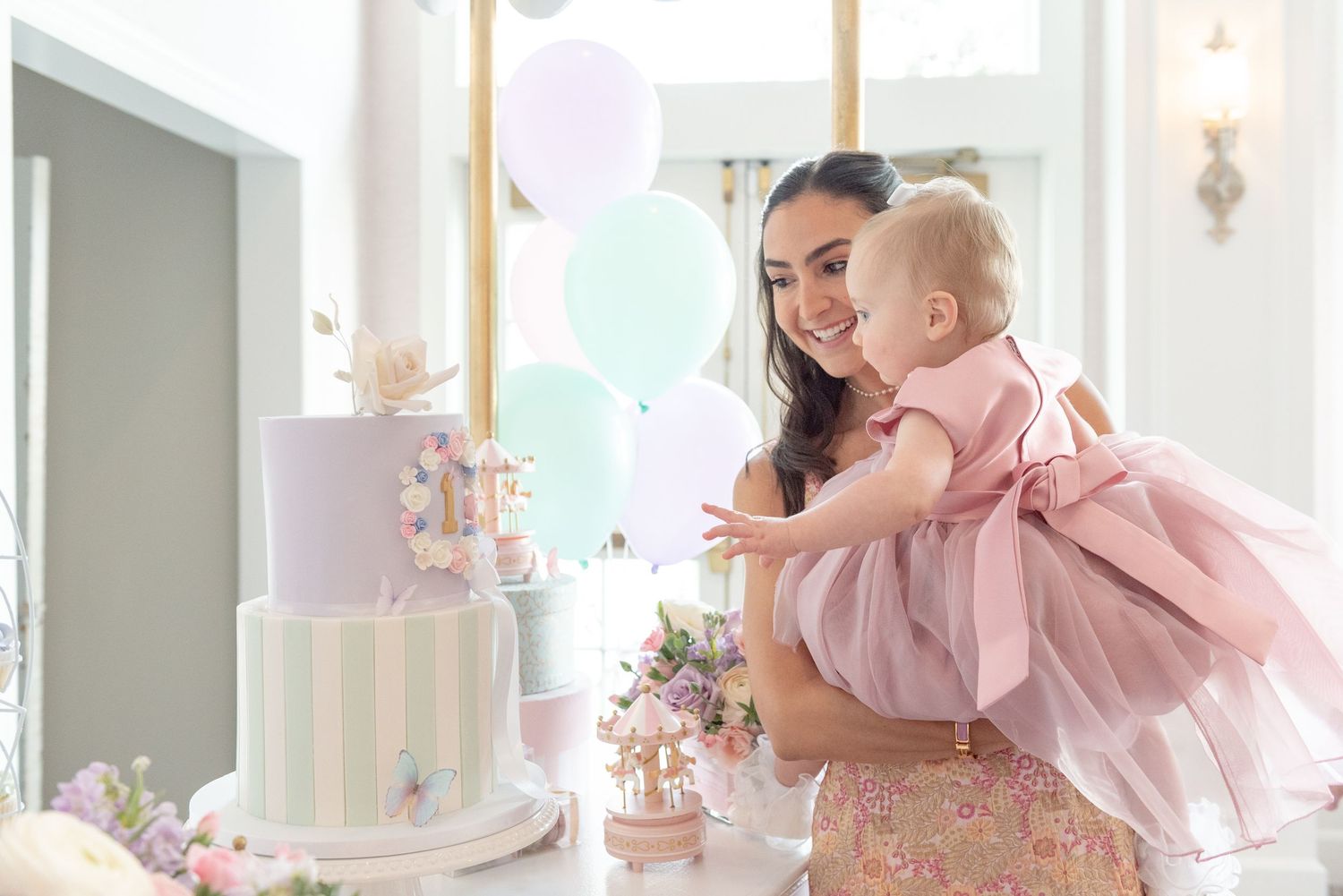 A baby in a pink dress plays with a parent in a bright, airy room decorated with pastel party decorations.
