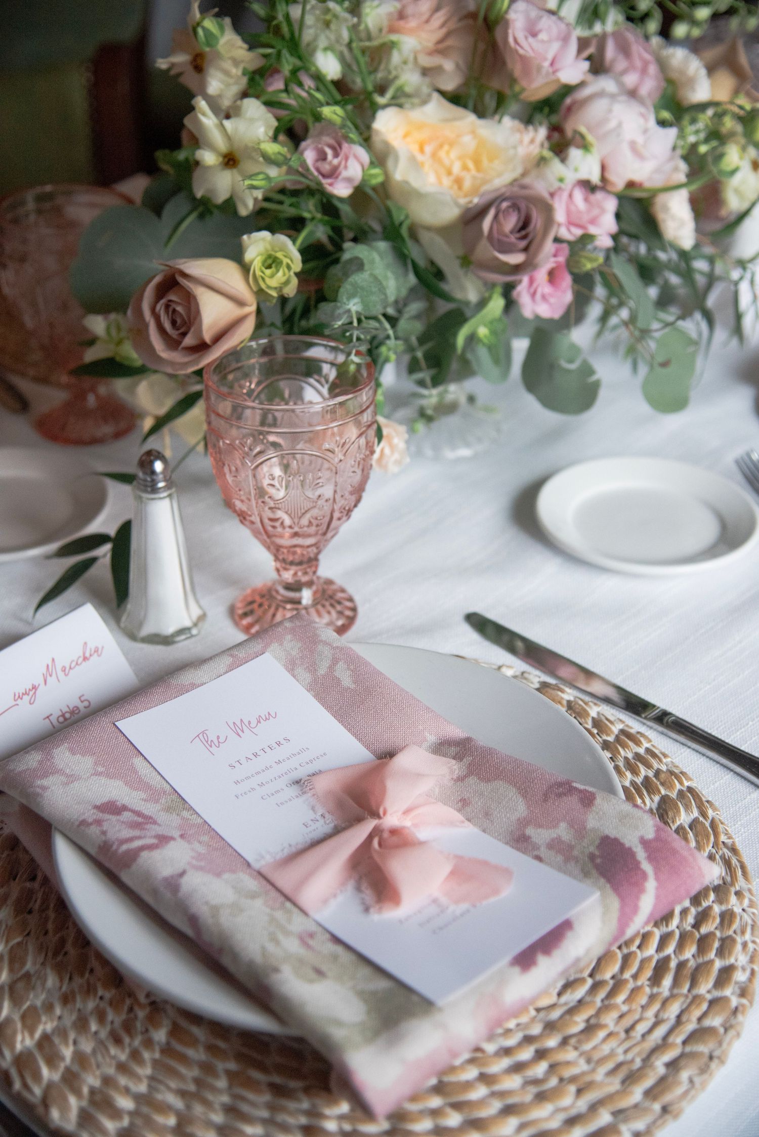 Elegant table setting with pink roses, vintage glassware, and delicate place cards adorned with blush ribbons on woven placemats.