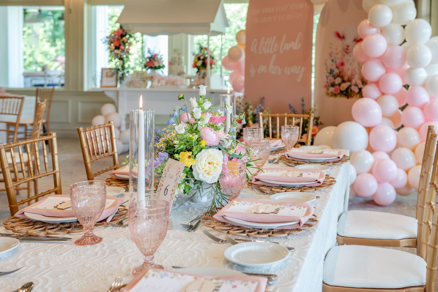 Pink and white balloon arch decorates an elegant baby shower table setting with gold chairs and floral centerpieces.