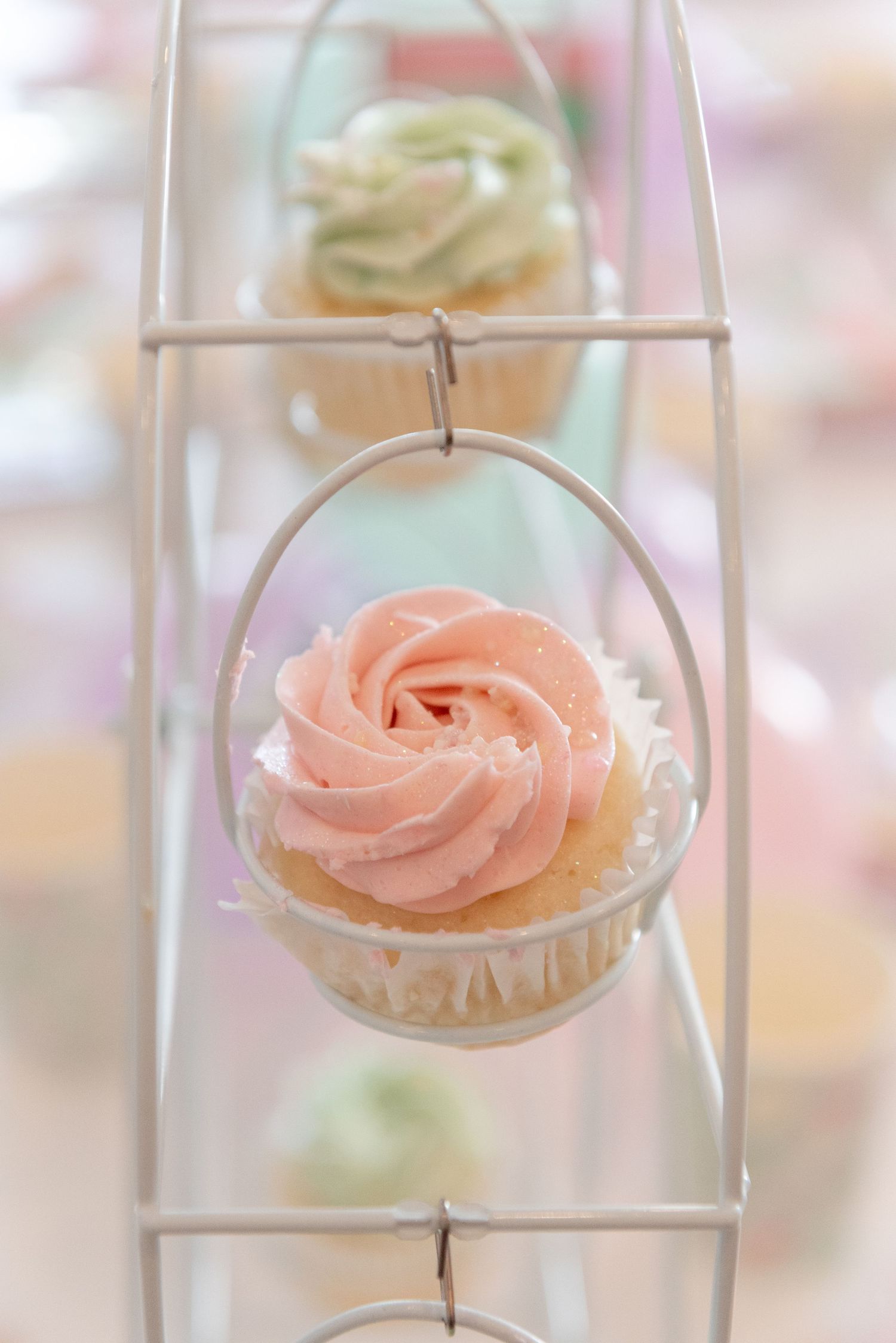 Pink frosted cupcakes with delicate rose swirl decorations displayed in a glass showcase with soft pastel background.