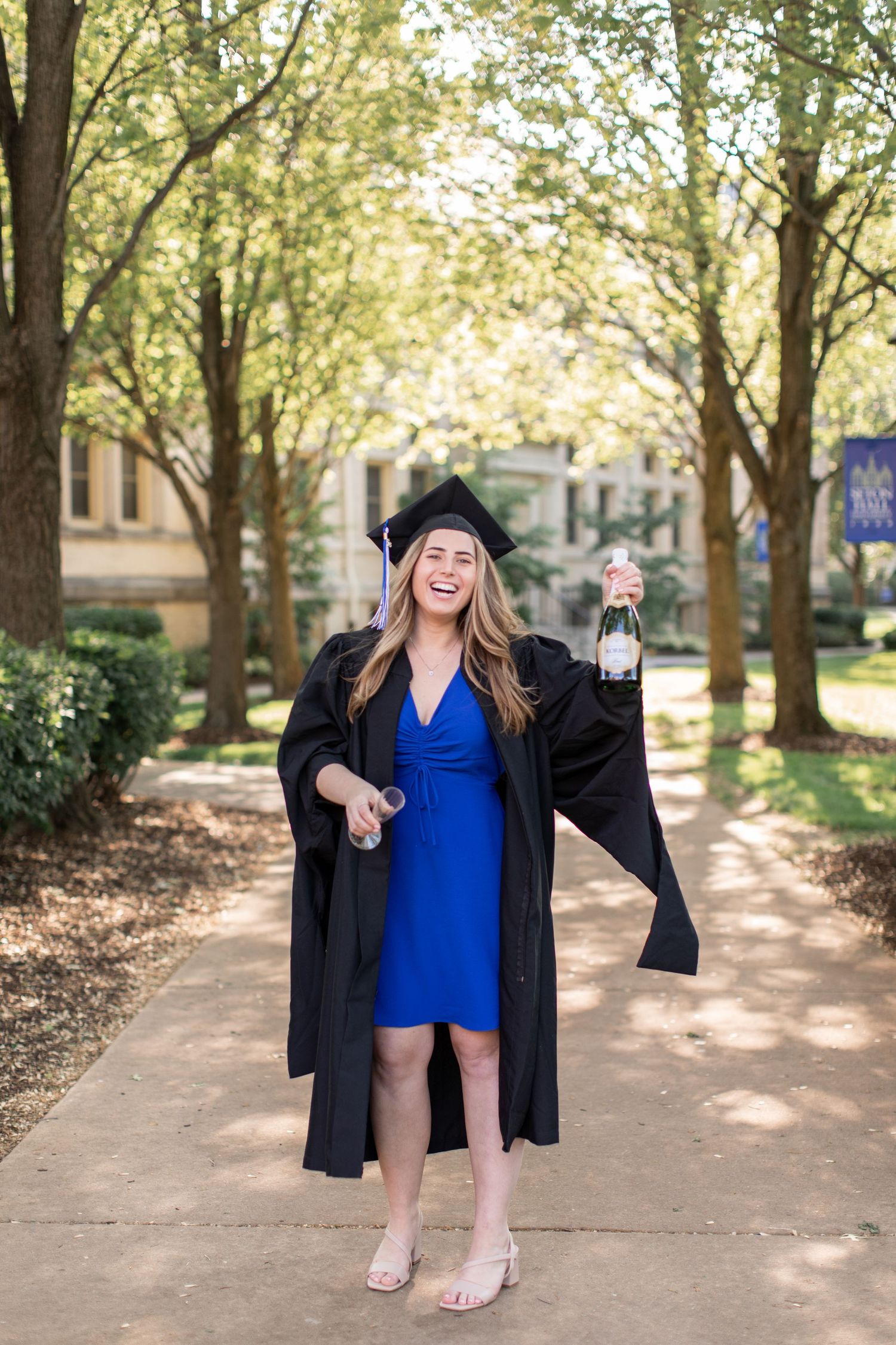 A graduate in a black cap and gown and blue dress poses joyfully on a tree-lined path during a sunny graduation photoshoot.