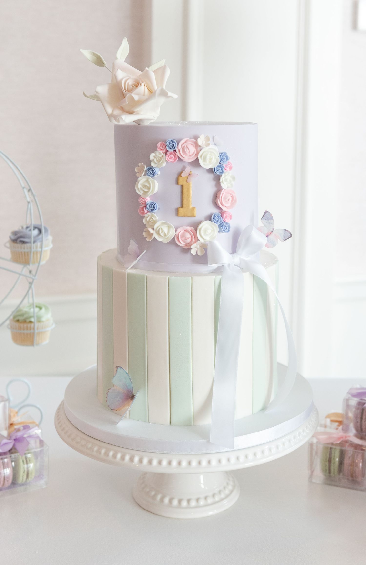 Elegant pastel-colored birthday cake with delicate floral decorations, butterflies, and mint green stripes on a white pedestal.