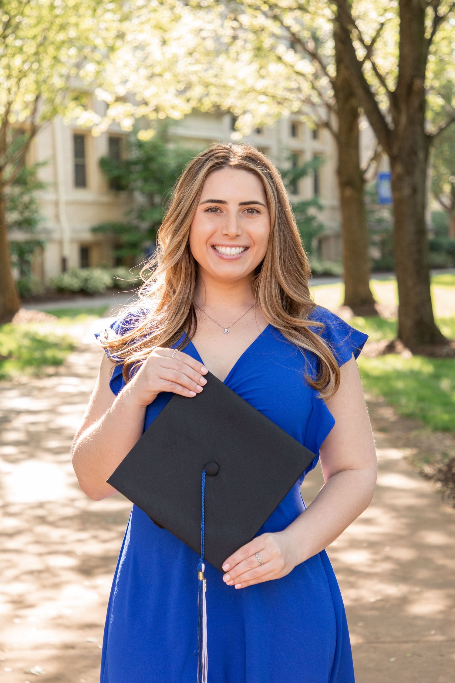 Graduate in royal blue dress holds up diploma and mortarboard cap while smiling outdoors on sunny campus grounds.
