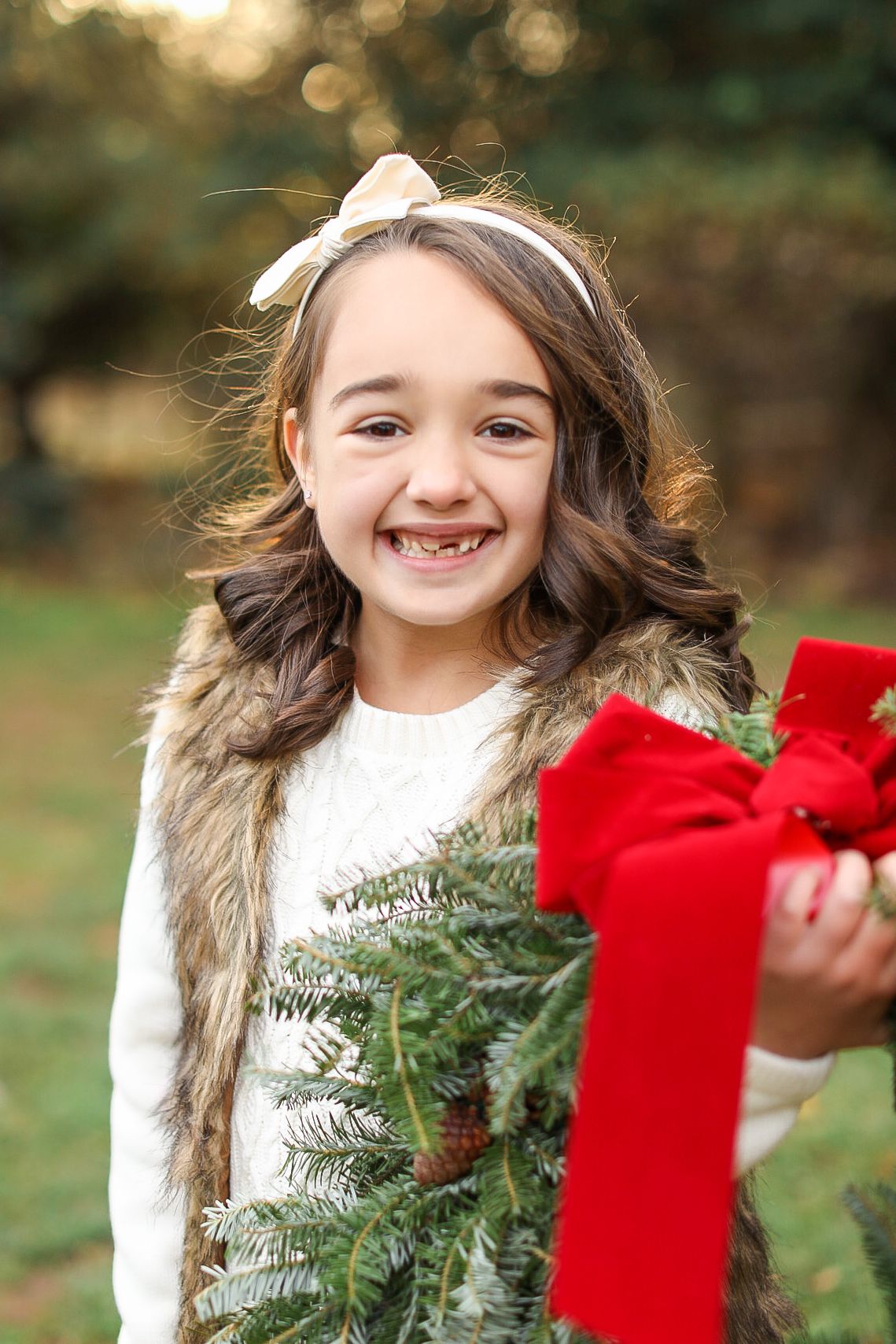 Child wearing fur vest and white sweater smiles while holding Christmas wreath with red bow outdoors.