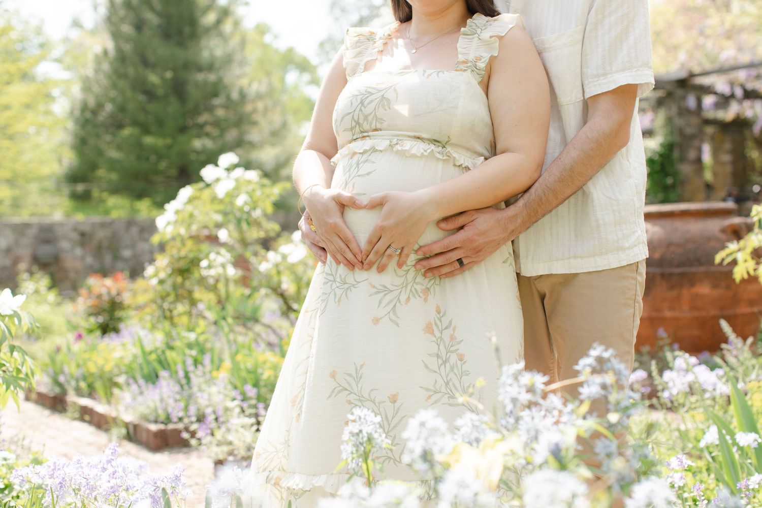 Expecting couple poses together in a garden setting with white wildflowers and natural greenery.