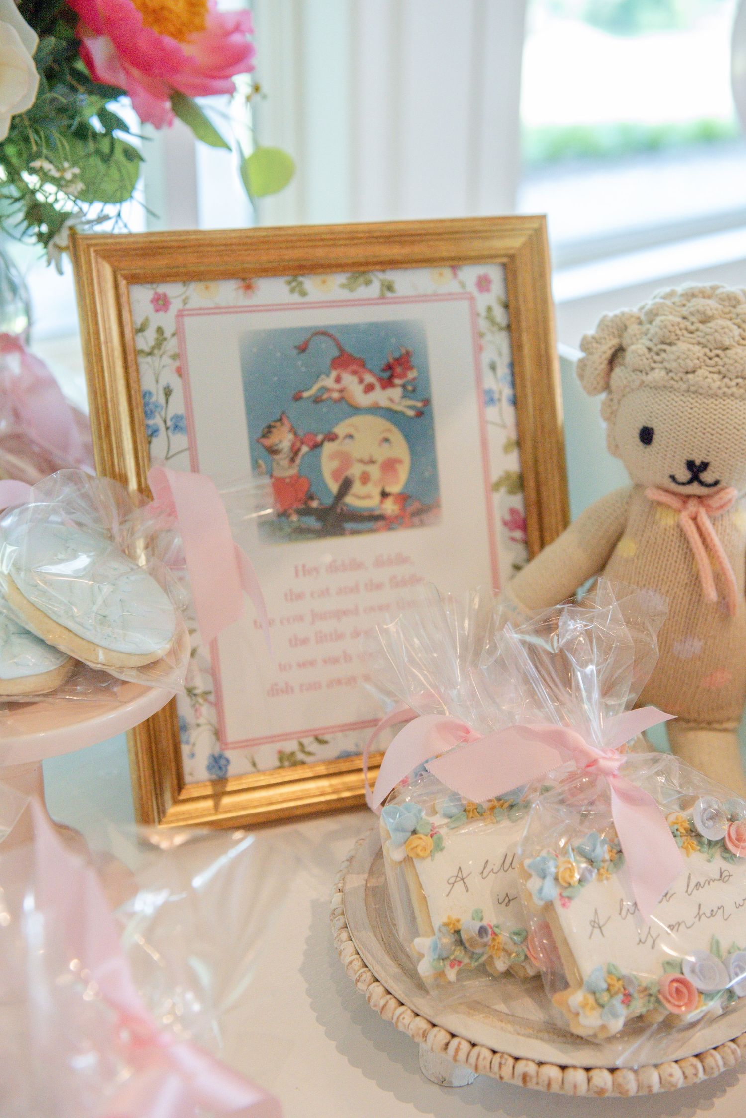Vintage-style nursery rhyme themed party decor with pink ribbons, framed illustrations and plush toys on display.