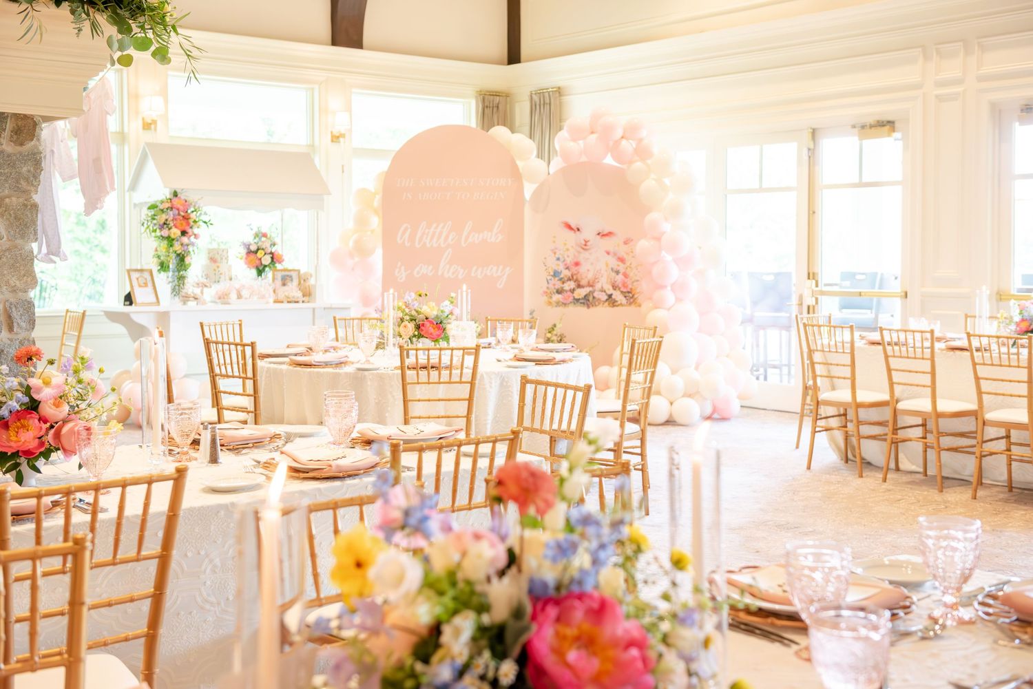 Elegant baby shower venue with pink decor, gold chiavari chairs, and colorful floral centerpieces in a bright room.