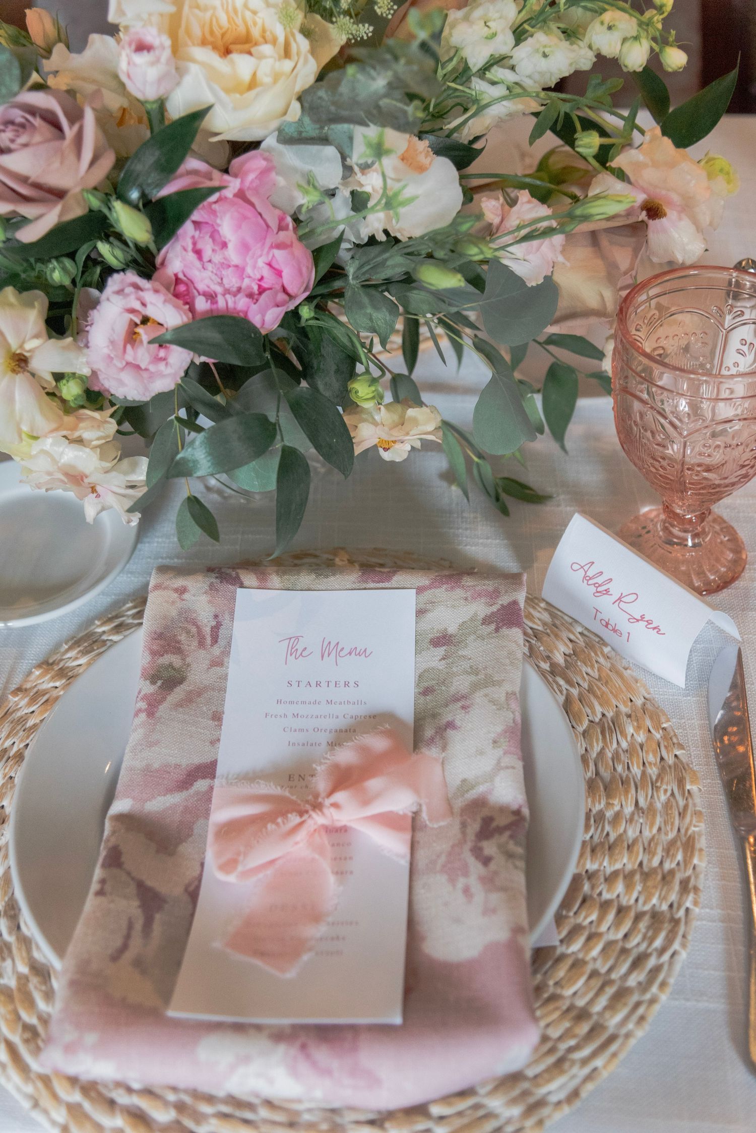 Elegant table setting with pink floral arrangements, woven placemats, and blush wedding invitations in romantic style.