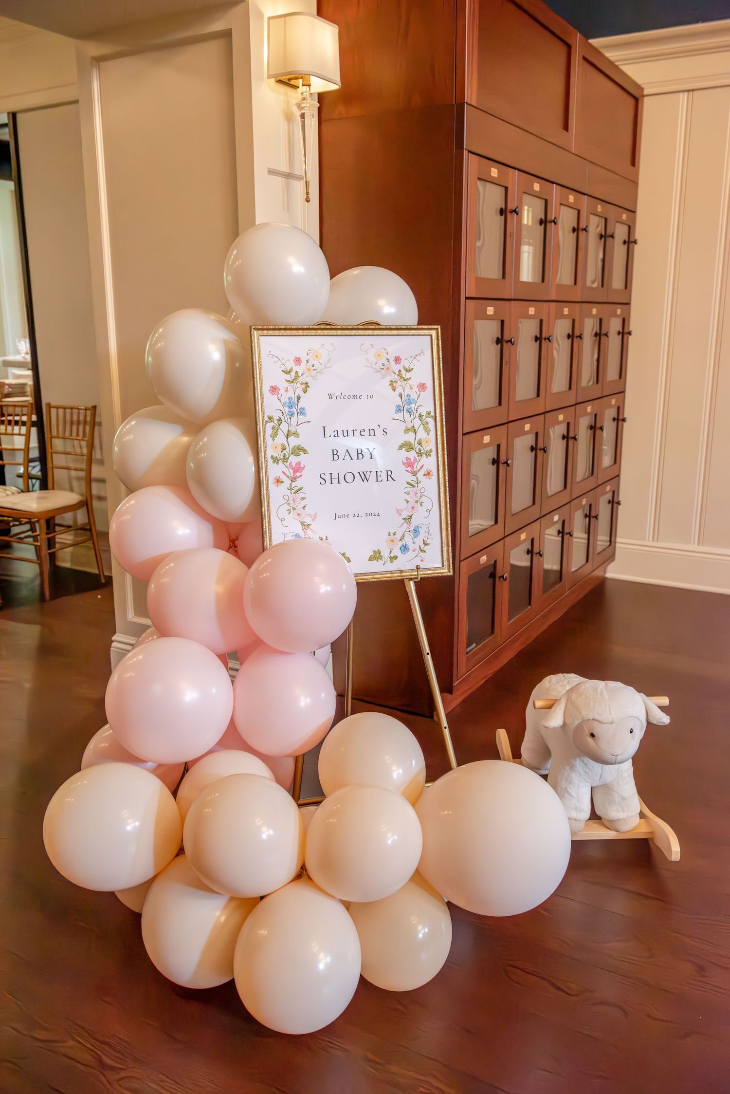 White and pink balloon arch and welcome sign decorations line a wooden hallway with elegant lighting.
