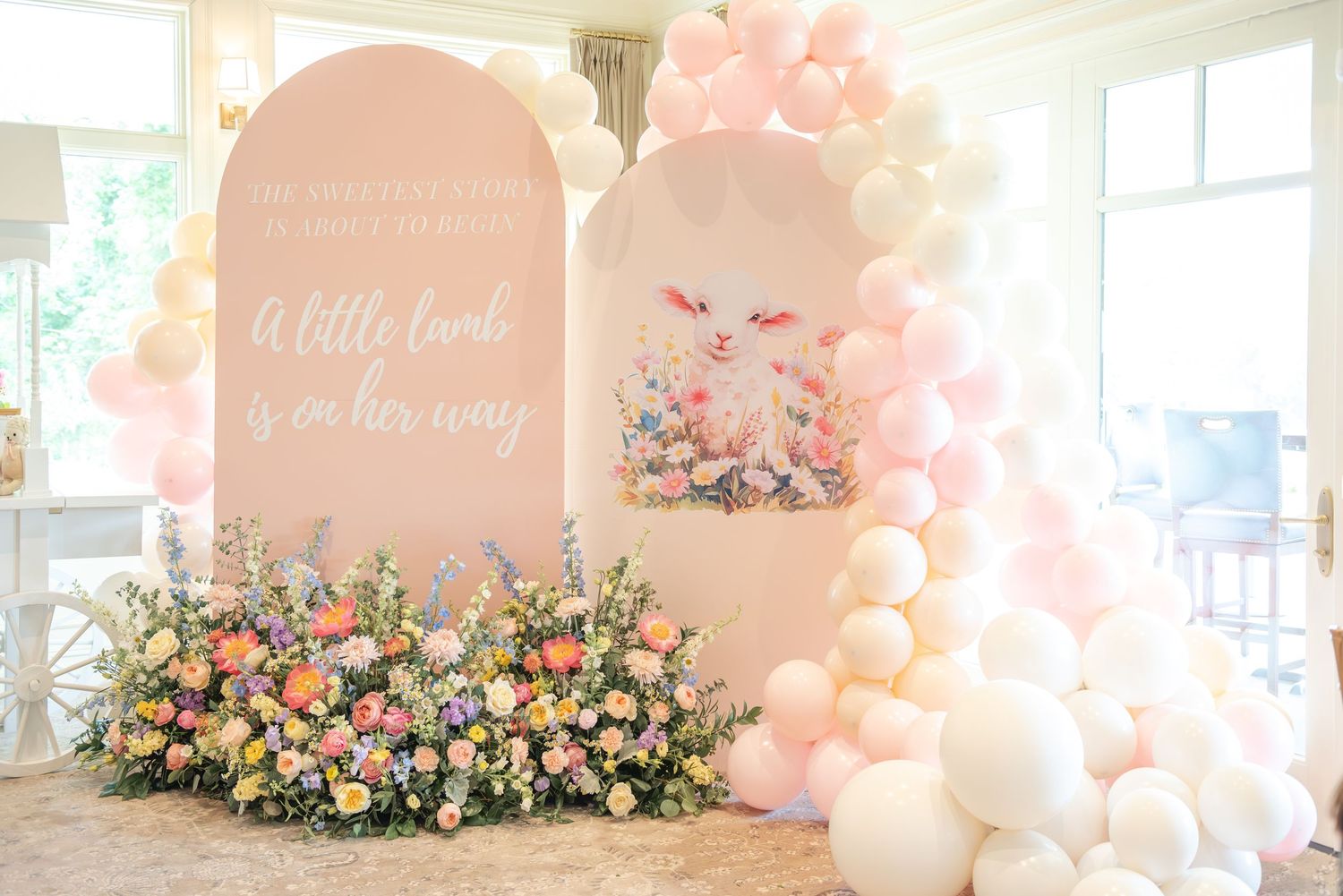Pink and white balloon arch with floral arrangements decorates a bright indoor baby shower celebration space.