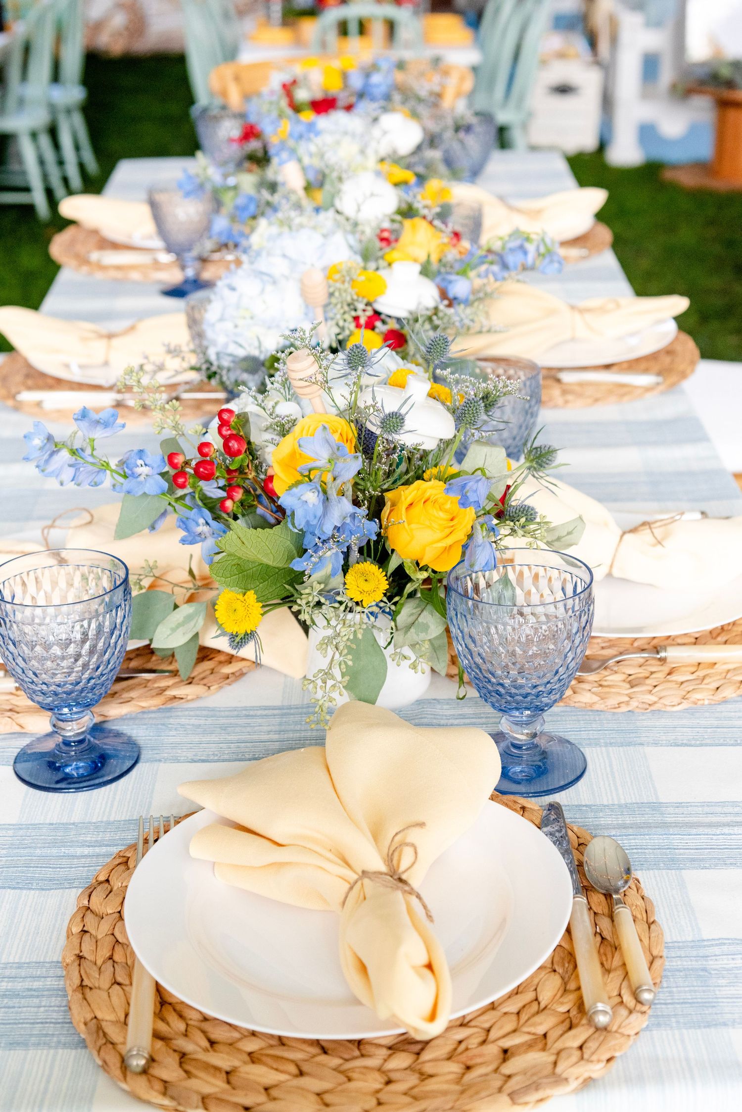 Elegant table setting with woven placemats, blue glassware, and colorful floral centerpieces on a white outdoor table.