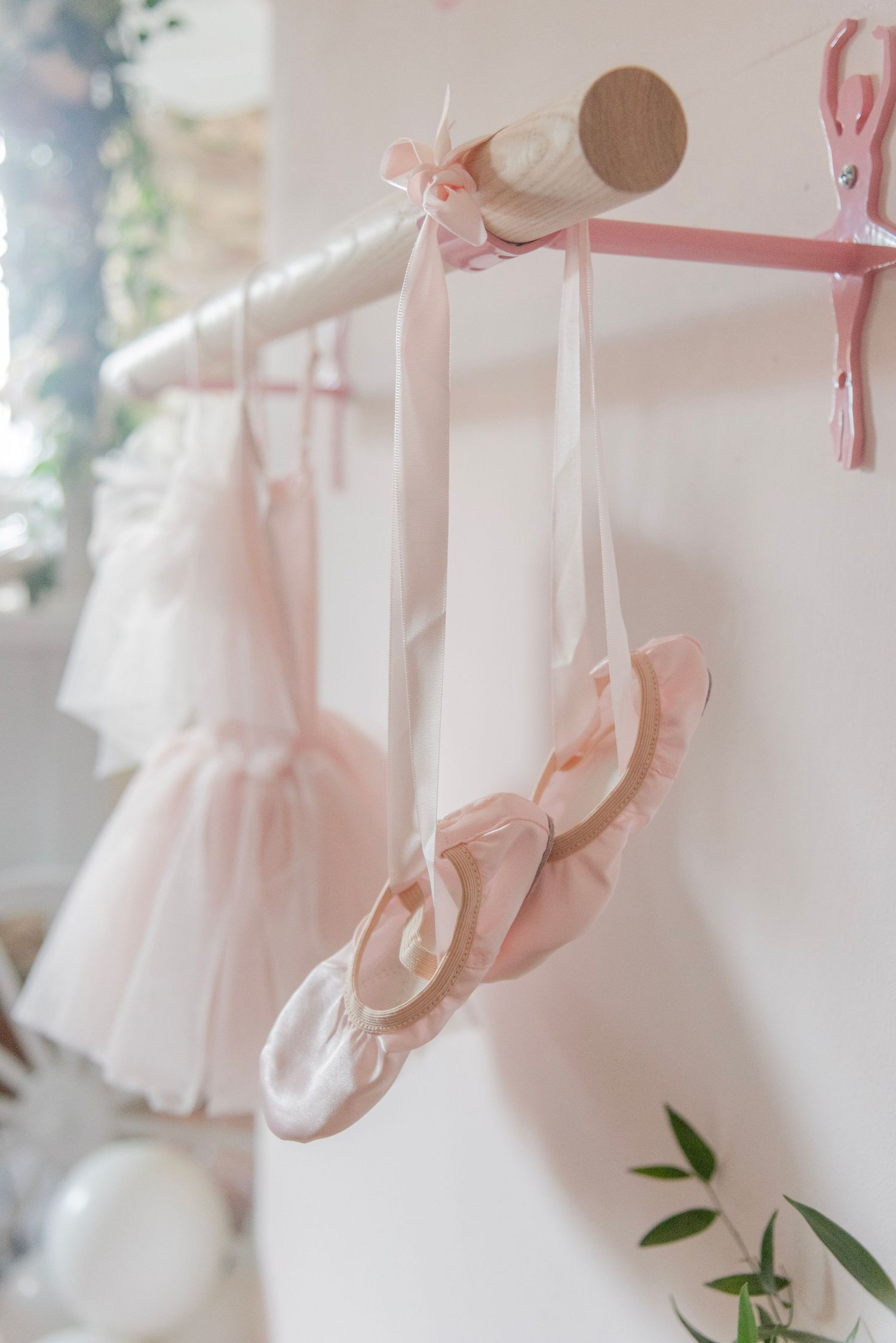 Delicate pink ballet pointe shoes and tutus hanging on display with soft lighting and green foliage accents.