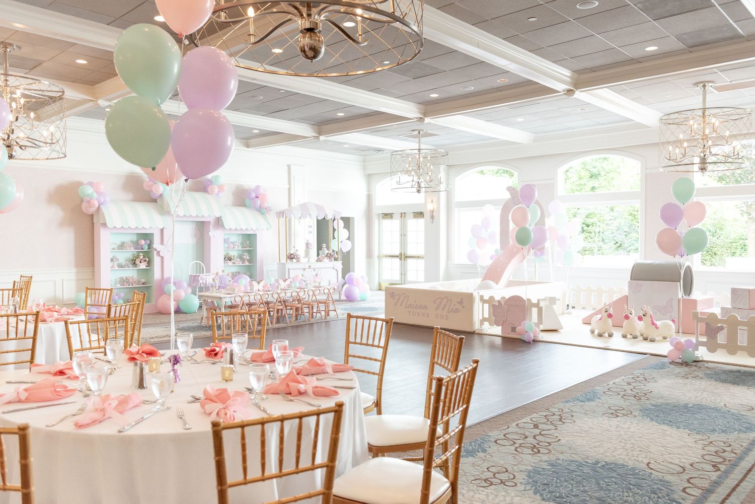 Elegant party venue with pink and pastel balloon decorations, round tables with white linens, and chiavari chairs.
