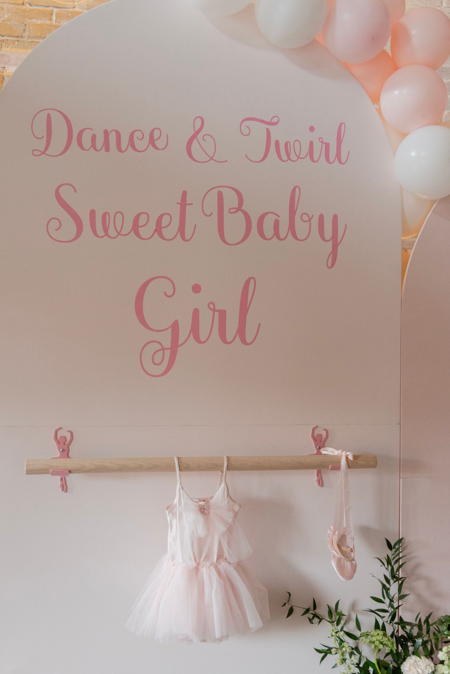 Pink and white ballet-themed baby shower decorations with tulle dresses, balloons, and greenery against a brick wall.