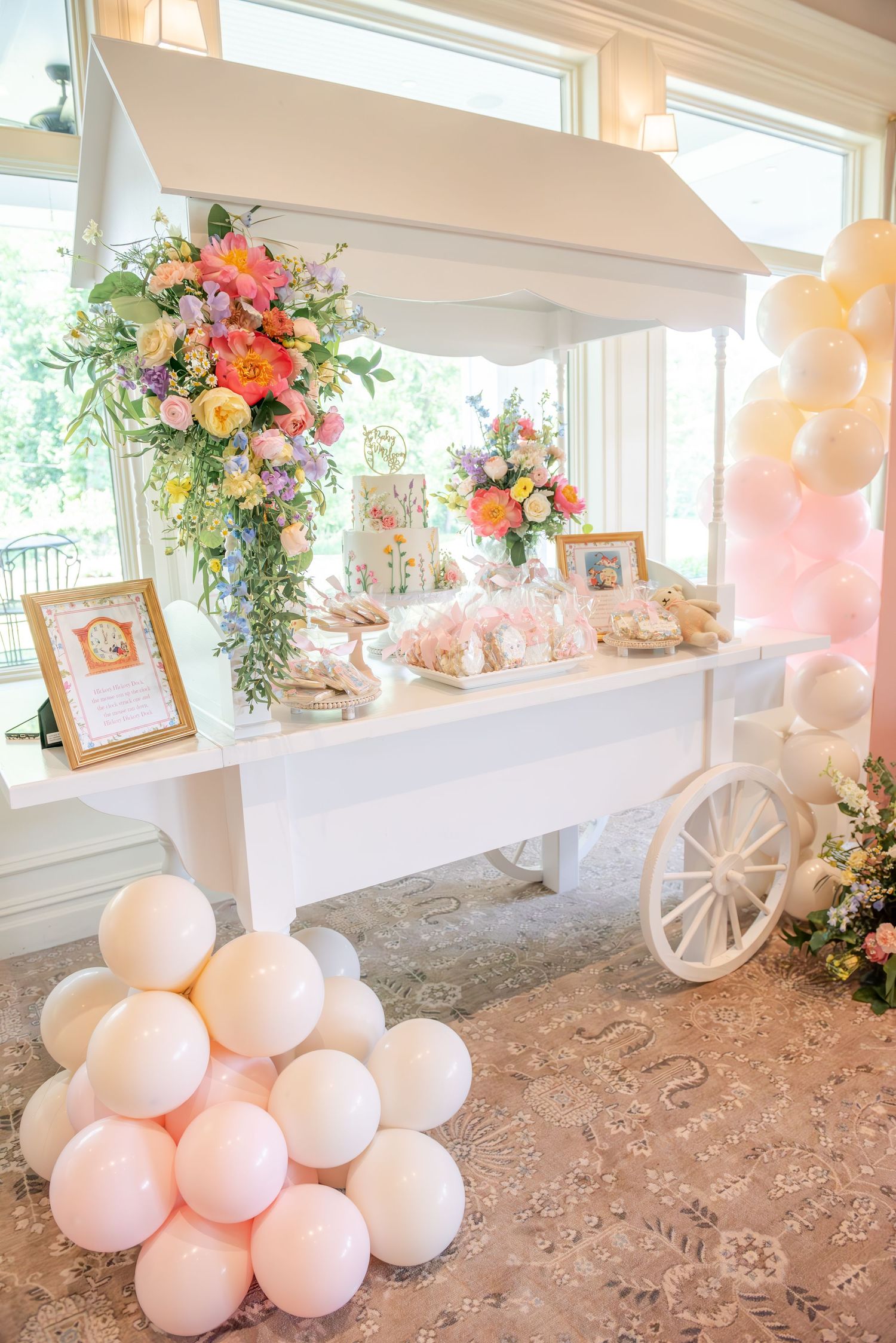 Elegant sweet cart display features white balloons, floral arrangements, and a vintage cart in a bright indoor venue.