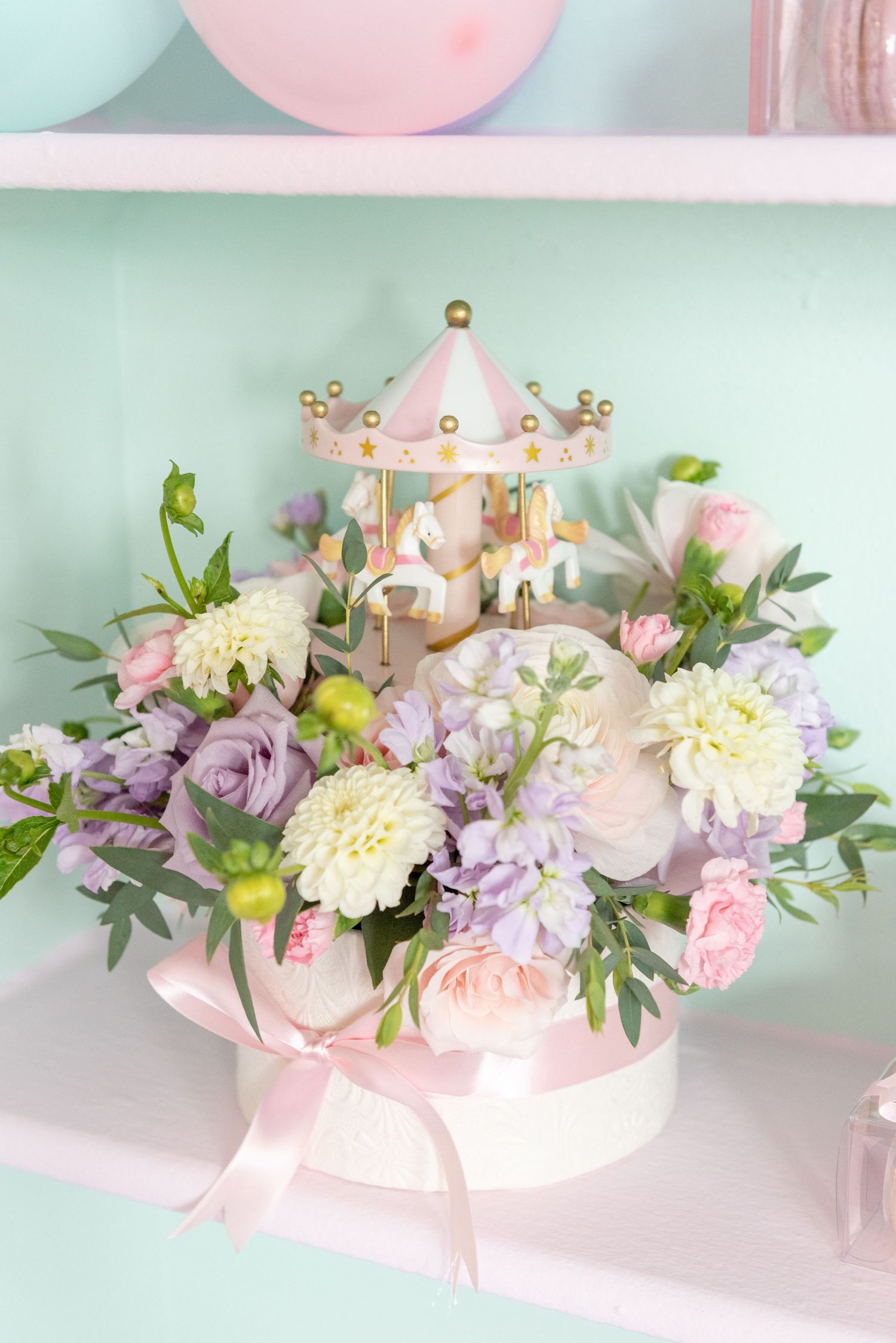 A delicate pink and purple floral arrangement with carnations and greenery in a soft pastel setting.