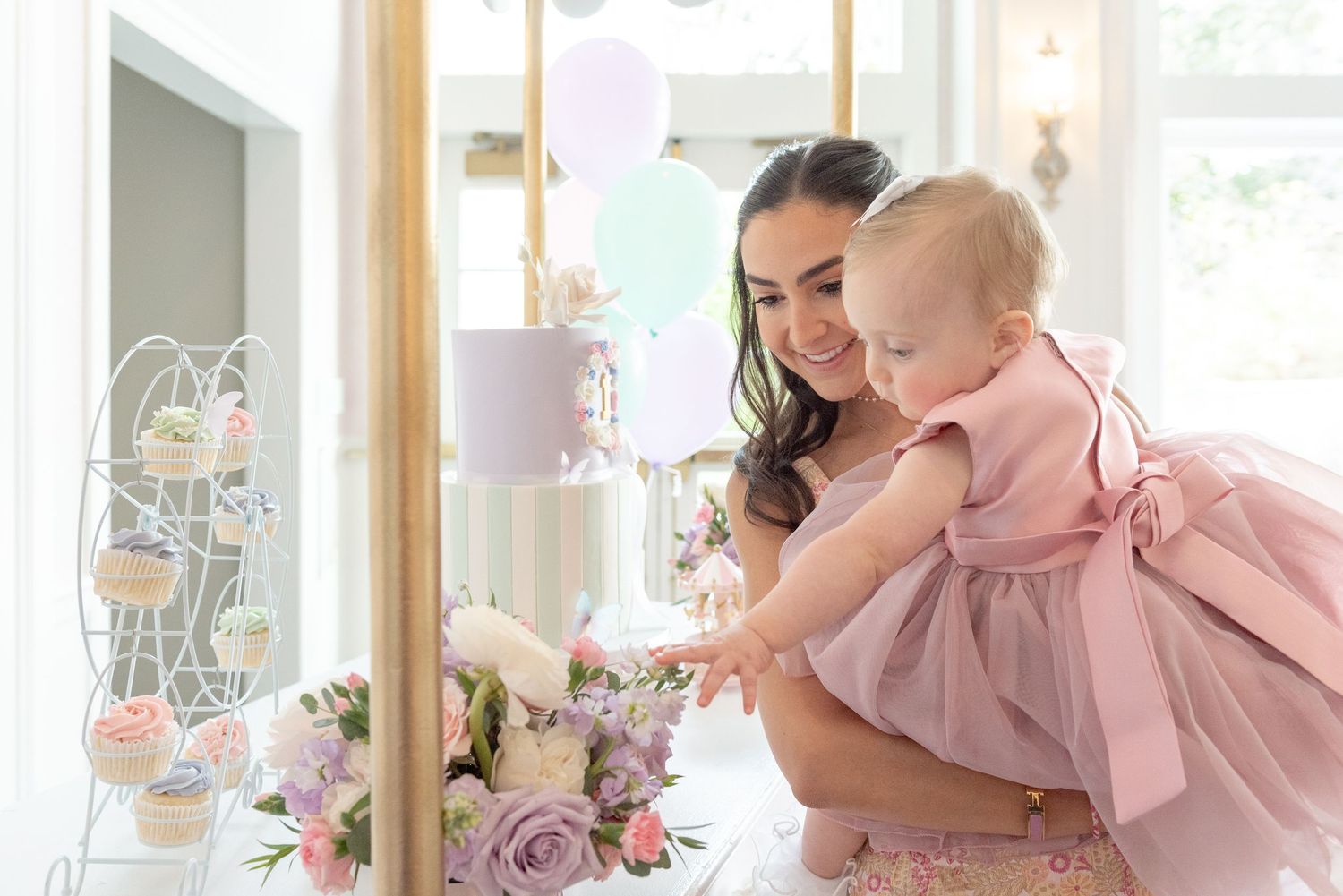A baby in a pink dress plays and smiles with someone in a light-colored dress against a bright interior background.
