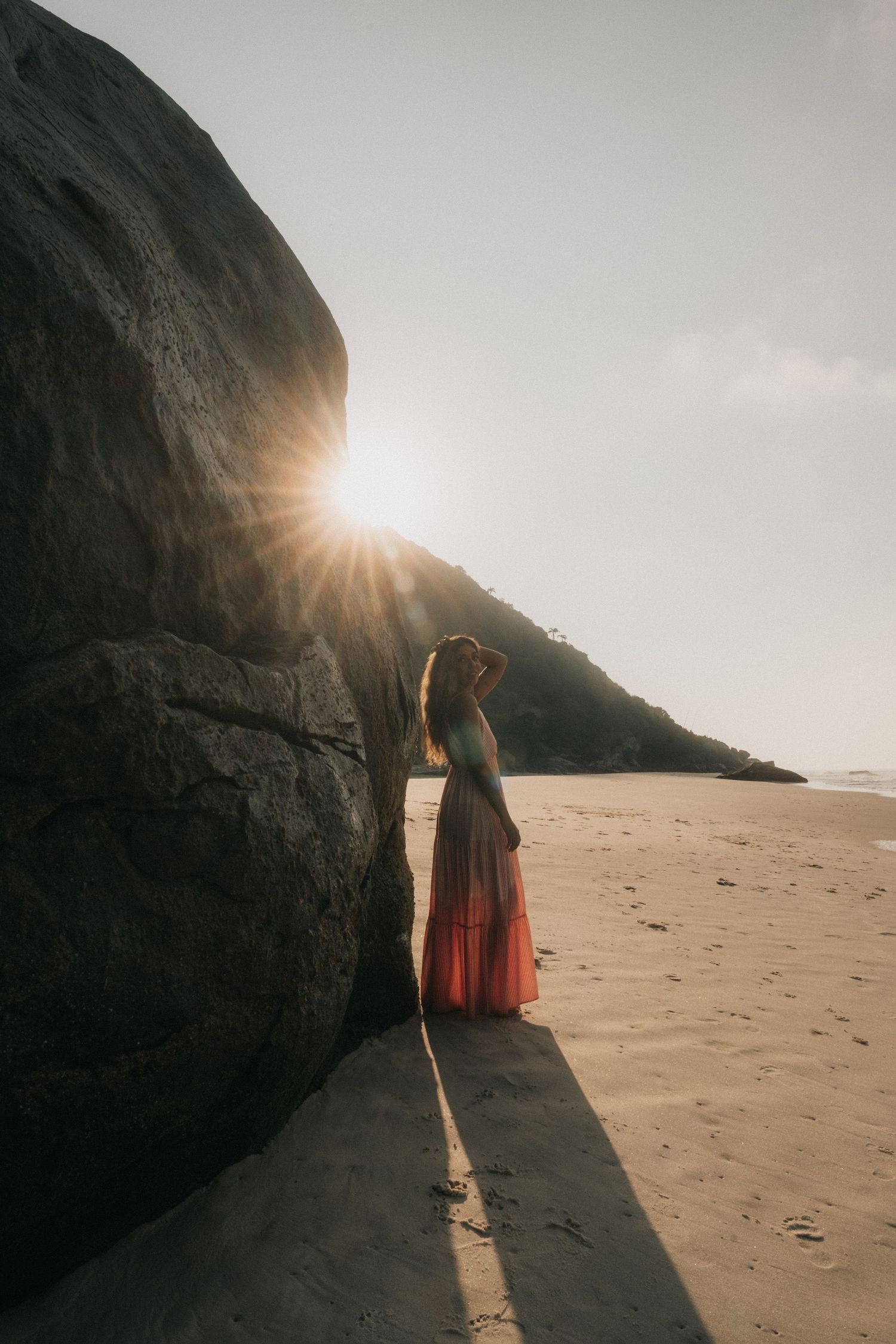 A person in red walks along a sandy beach at sunset while casting long shadows against dramatic coastal cliffs.