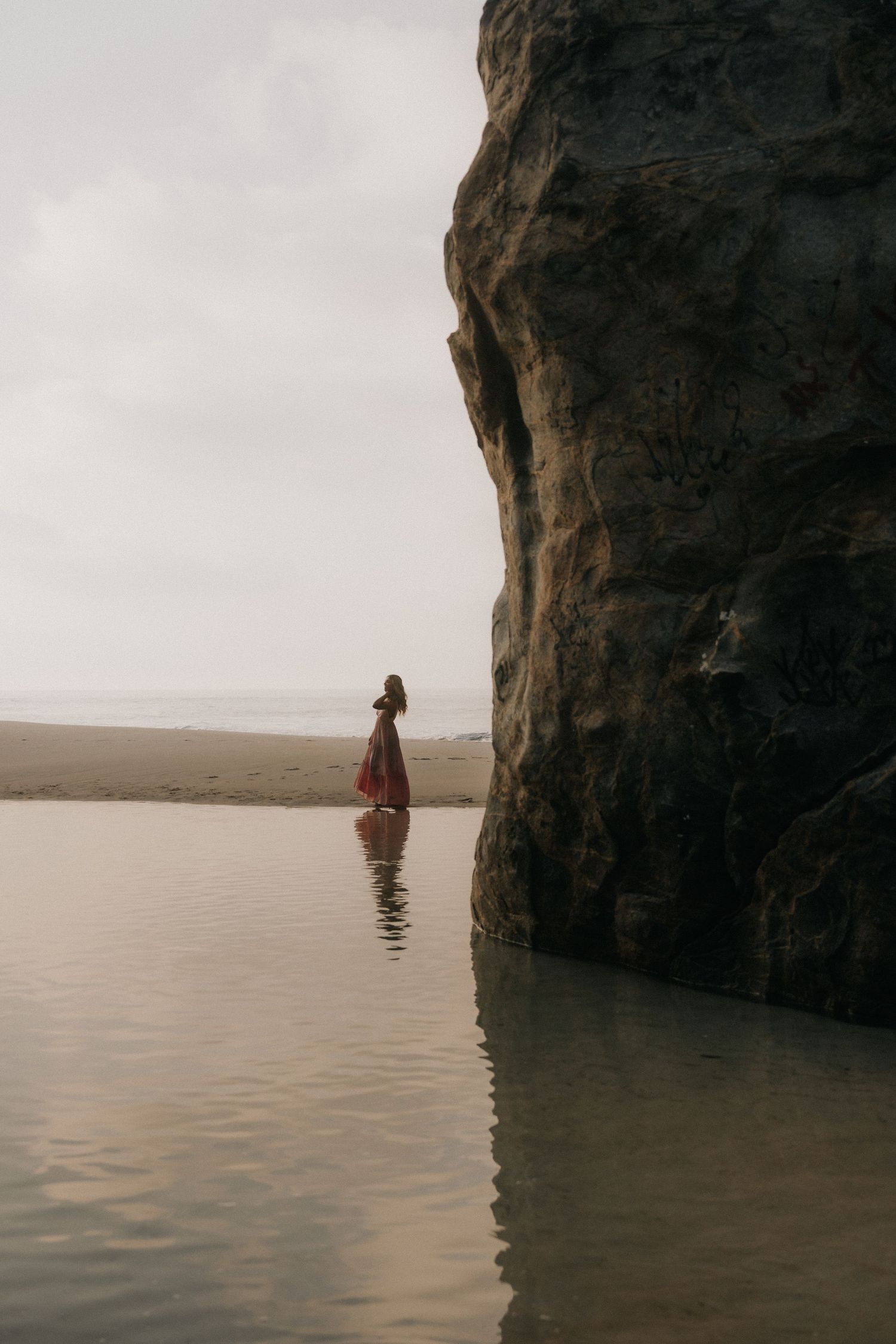 A silhouette of a figure in a dress stands in tranquil water beneath a towering coastal cliff face during sunset.