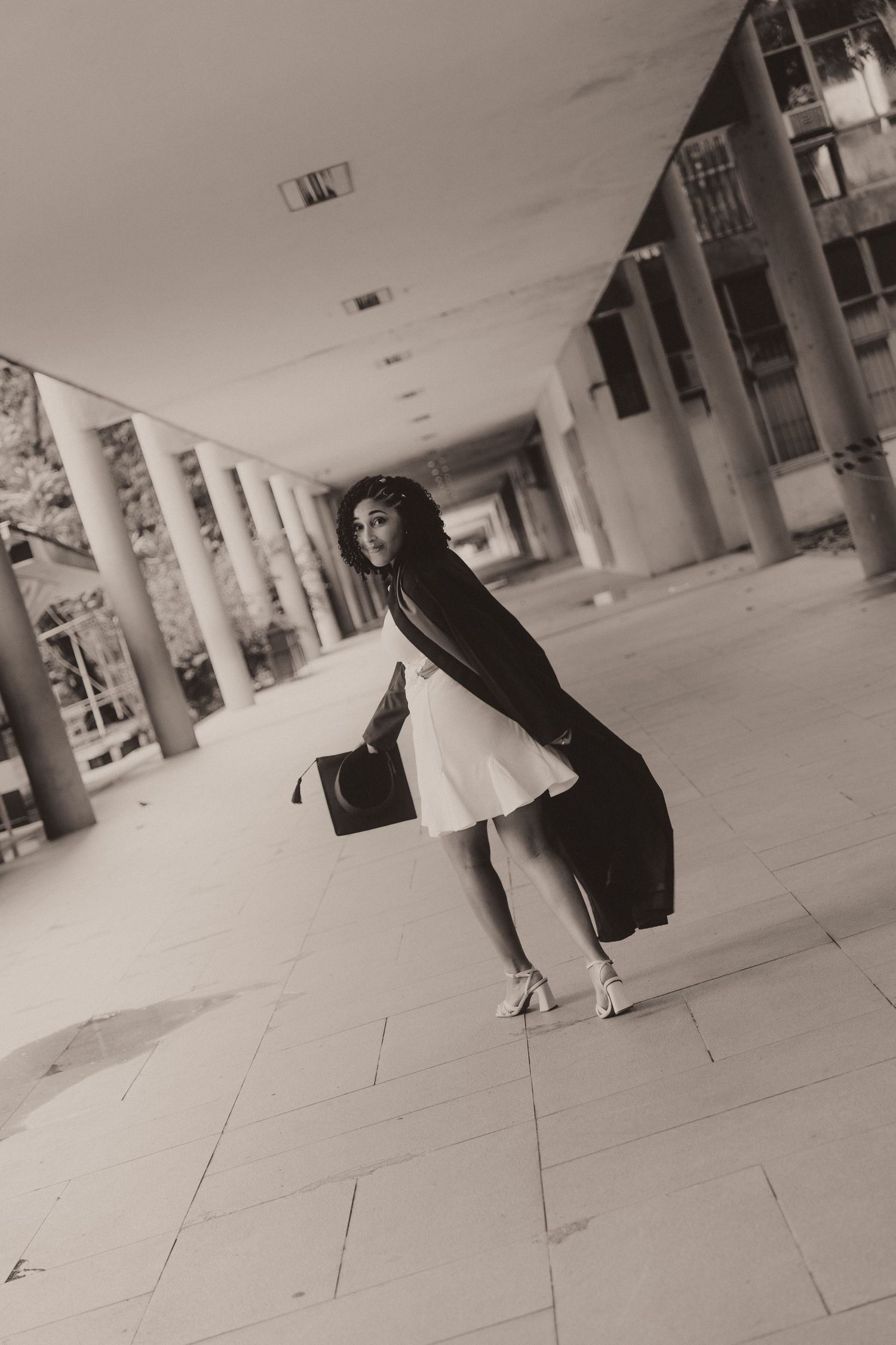 Figure in black dress and boots stands in repeating architectural walkway with white columns and tiled floor creating depth.