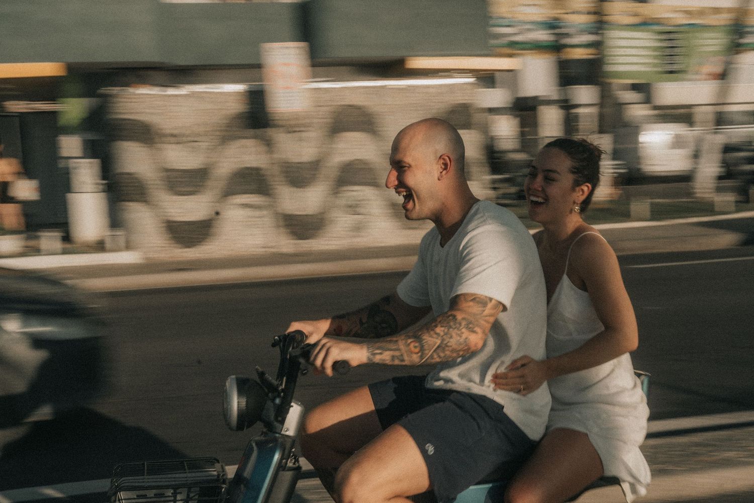 A couple rides a motorcycle along a street with a blurred sidewalk mural in the background.