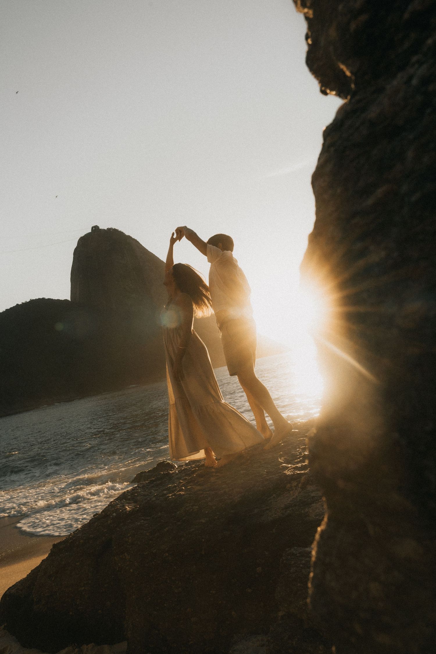 A romantic silhouette scene captured at sunset on a rocky beach with dramatic lens flare filtering through the cliffside.
