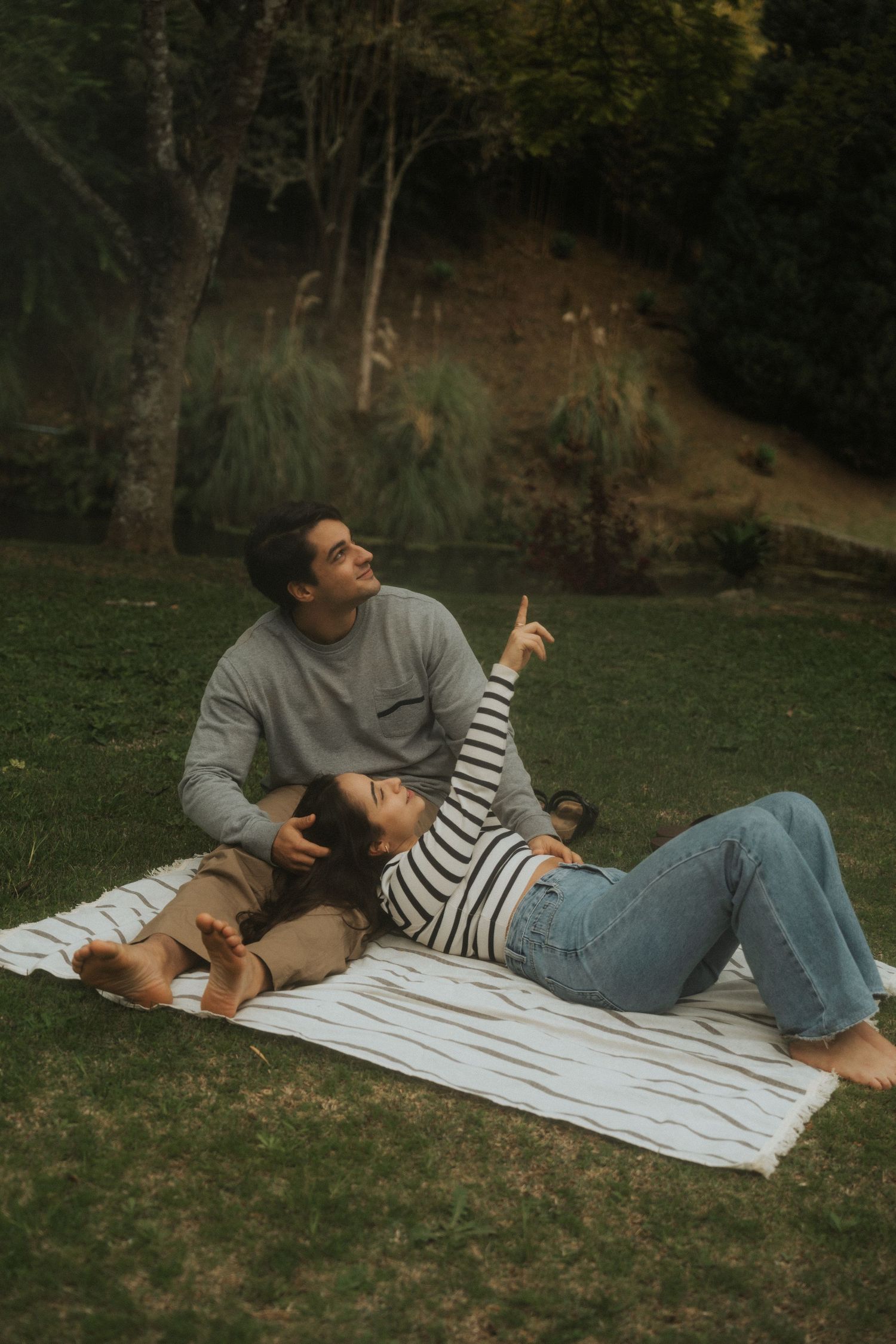 A couple relaxes together on a white blanket laid out on grass in a serene outdoor setting.