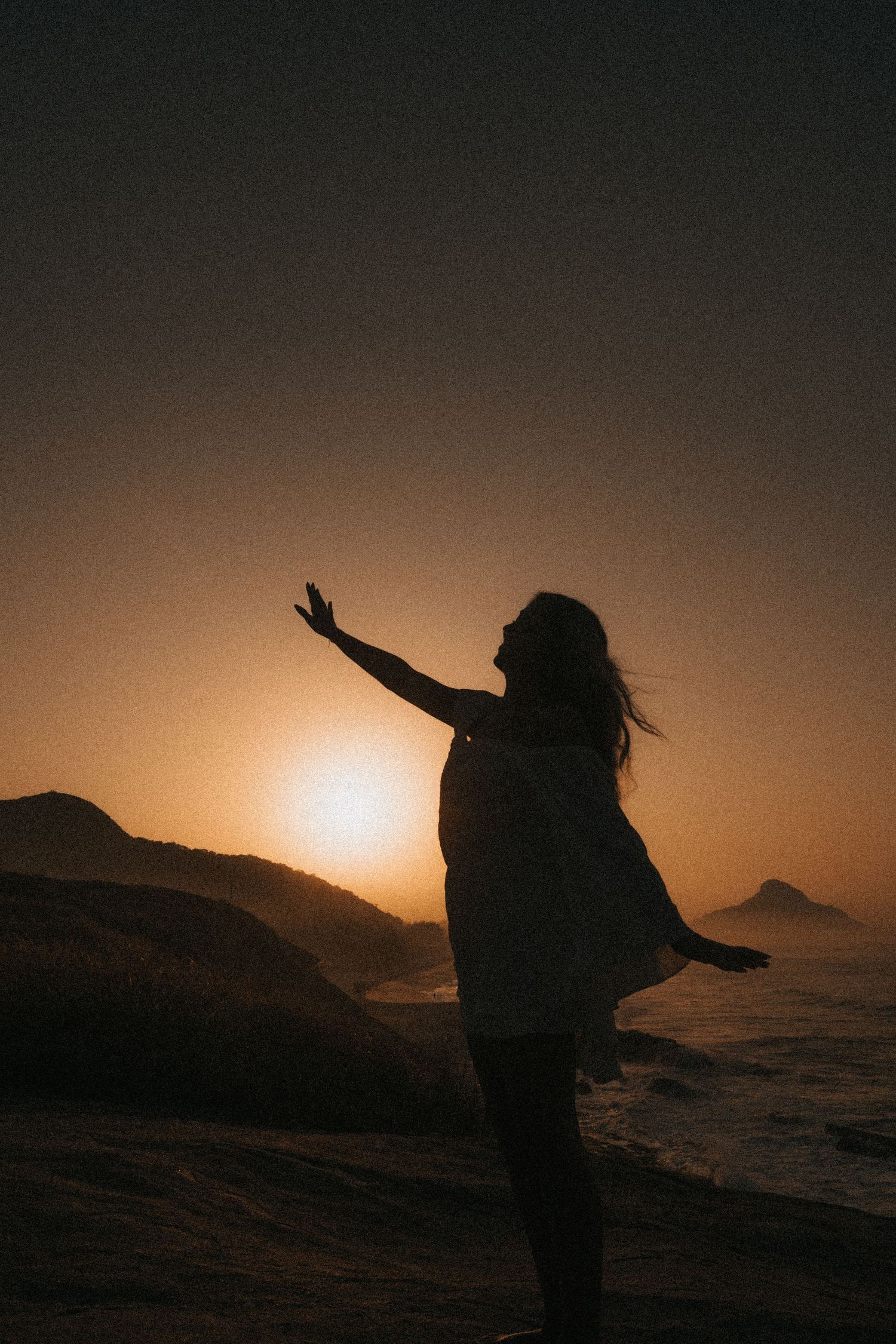 Silhouetted figure with outstretched arms stands against dramatic sunset over mountains and ocean waves.