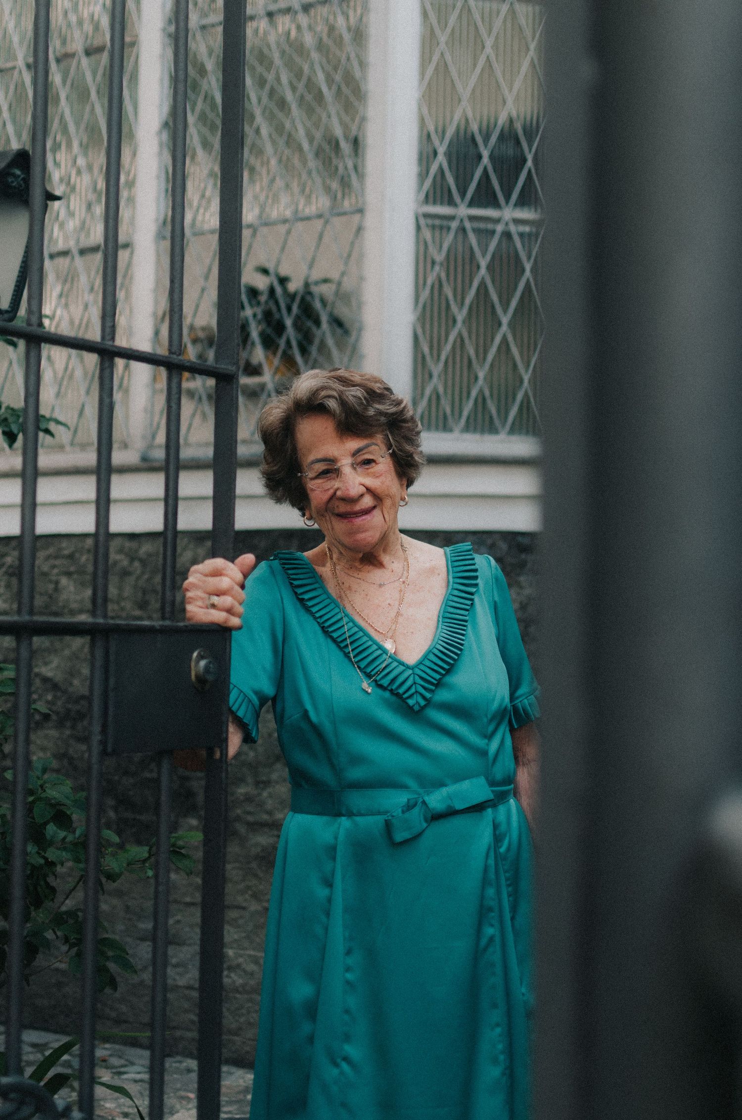 Person in a turquoise dress smiles and poses outdoors against a window with diamond-patterned grille.