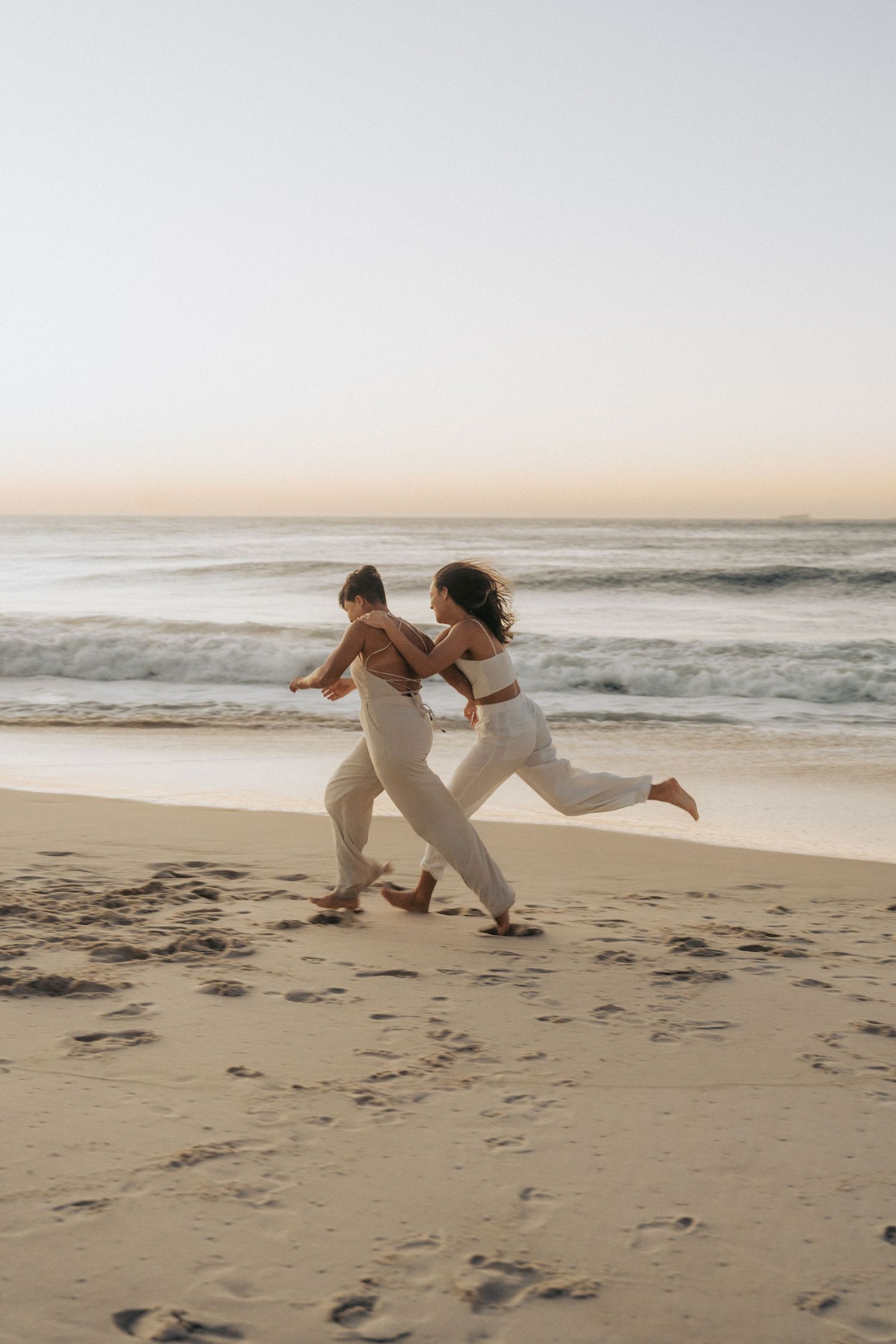 Two athletic women in light workout attire run joyfully along a misty beach at sunset.