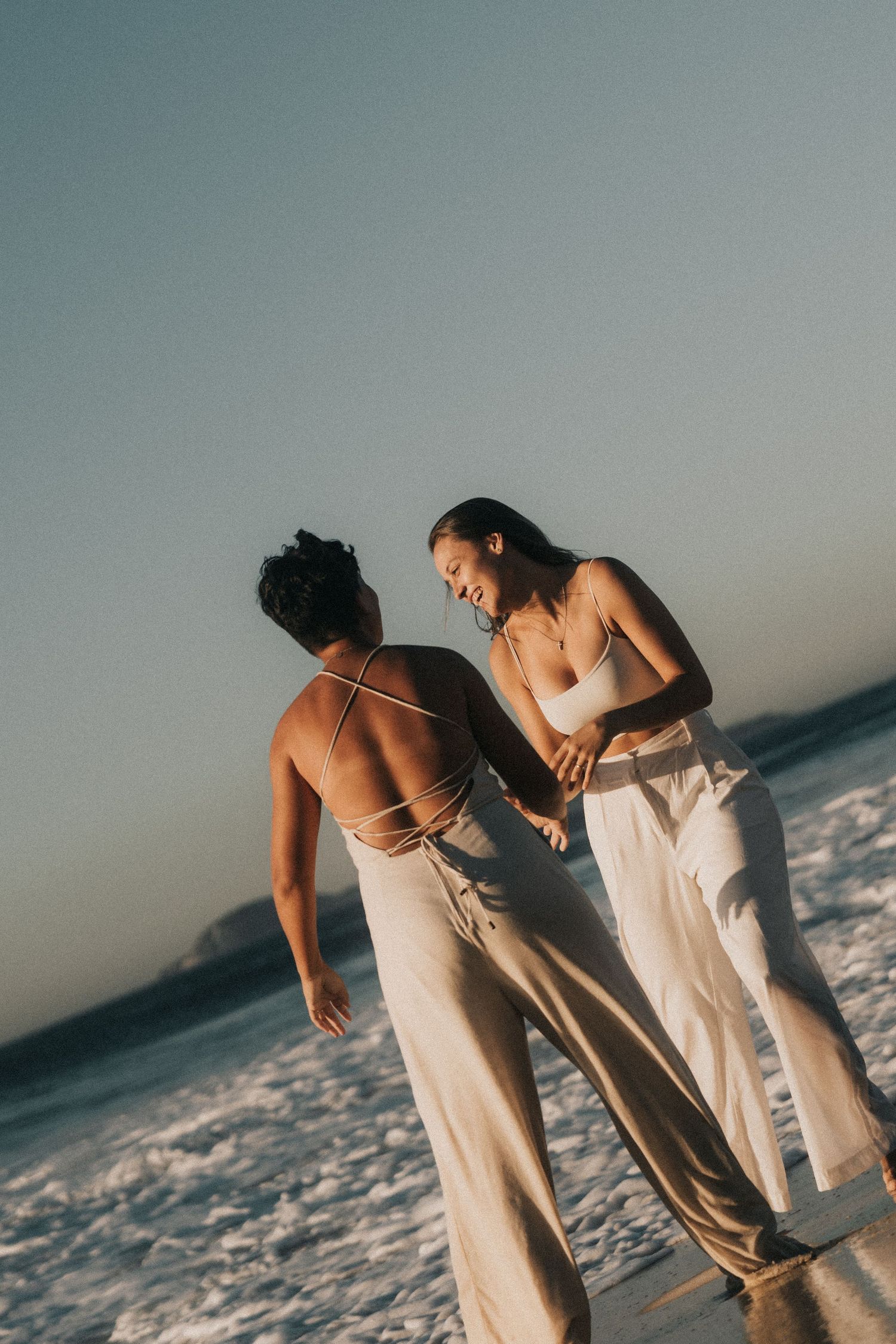 Two people in beige outfits sharing a playful moment while walking on a beach at sunset.