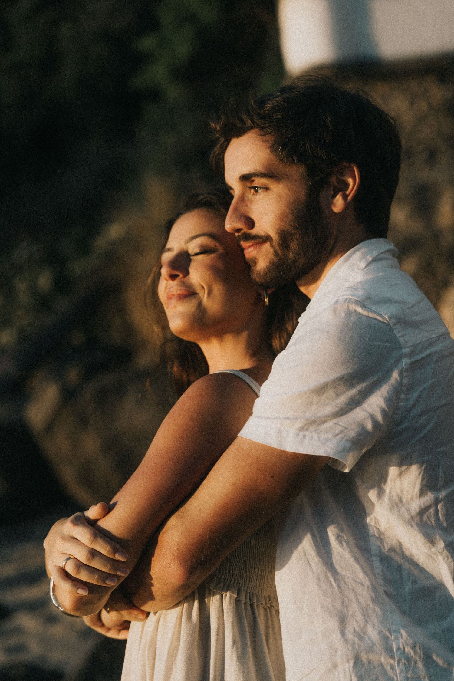 A tender romantic moment captured at sunset as a couple embraces while wearing white clothing against a warm golden backdrop.
