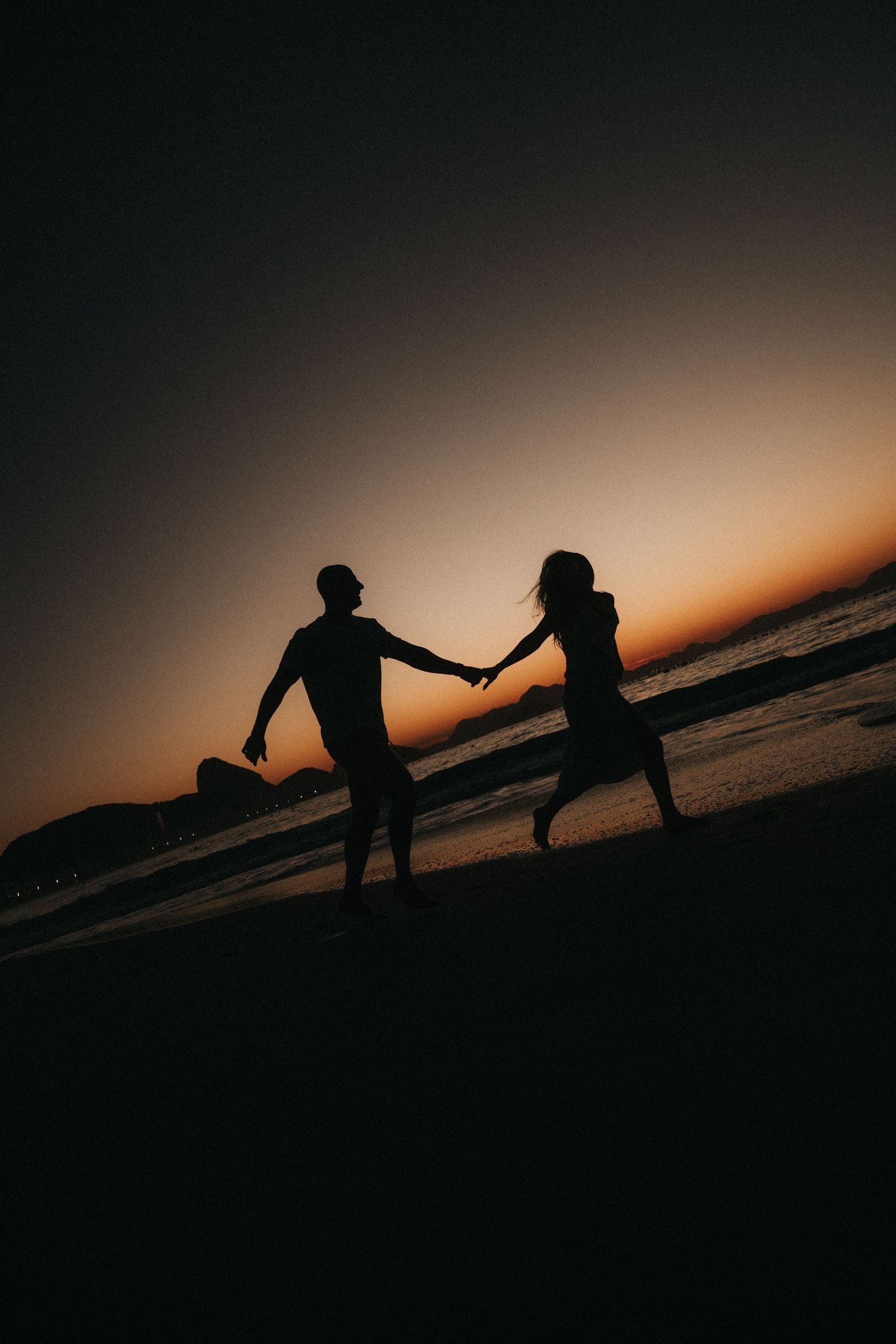 Silhouetted couple holding hands on beach against dramatic sunset sky backdrop.