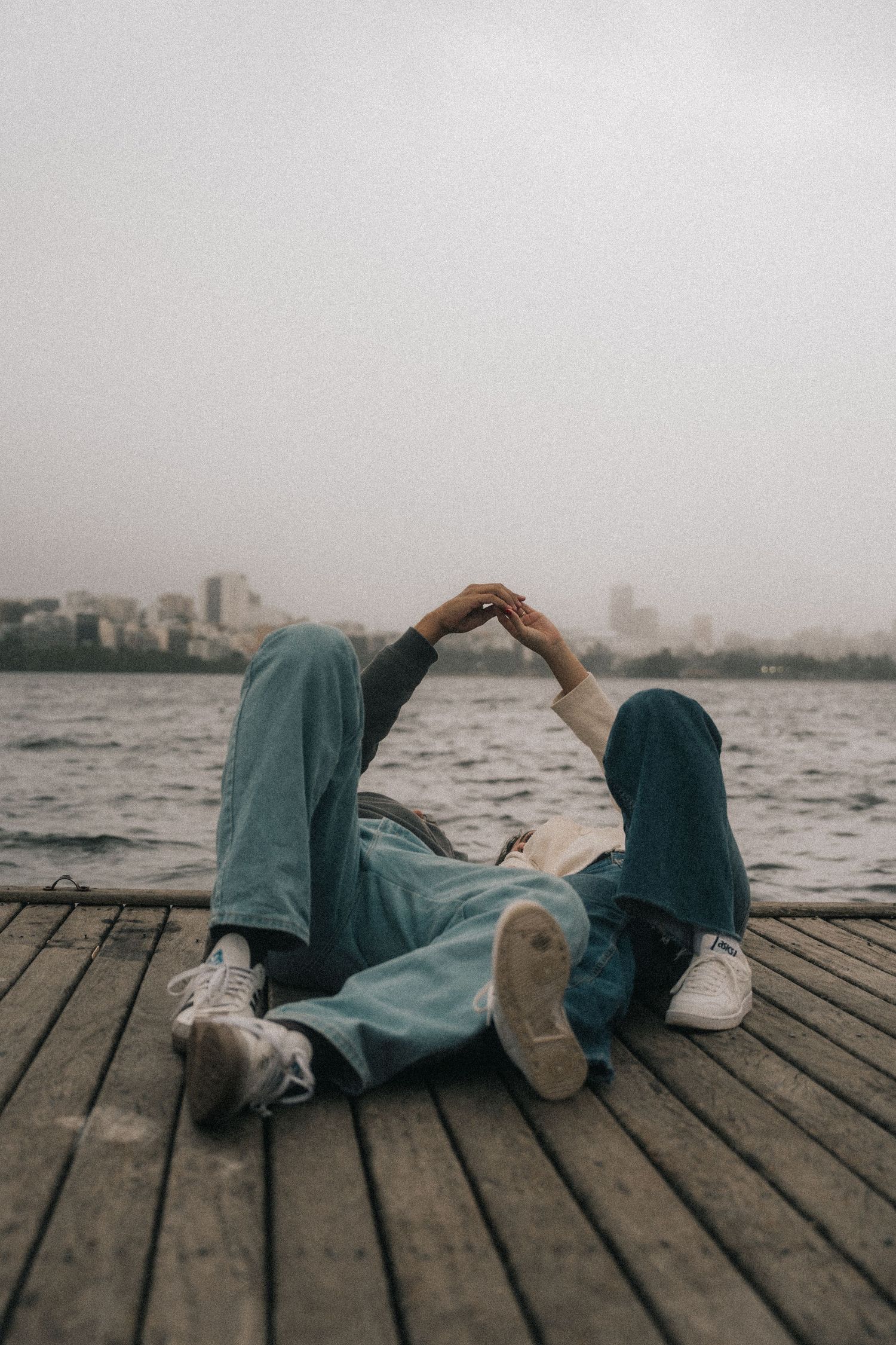A person lies on a wooden dock wearing a denim jacket and white sneakers against a misty city skyline across the water.