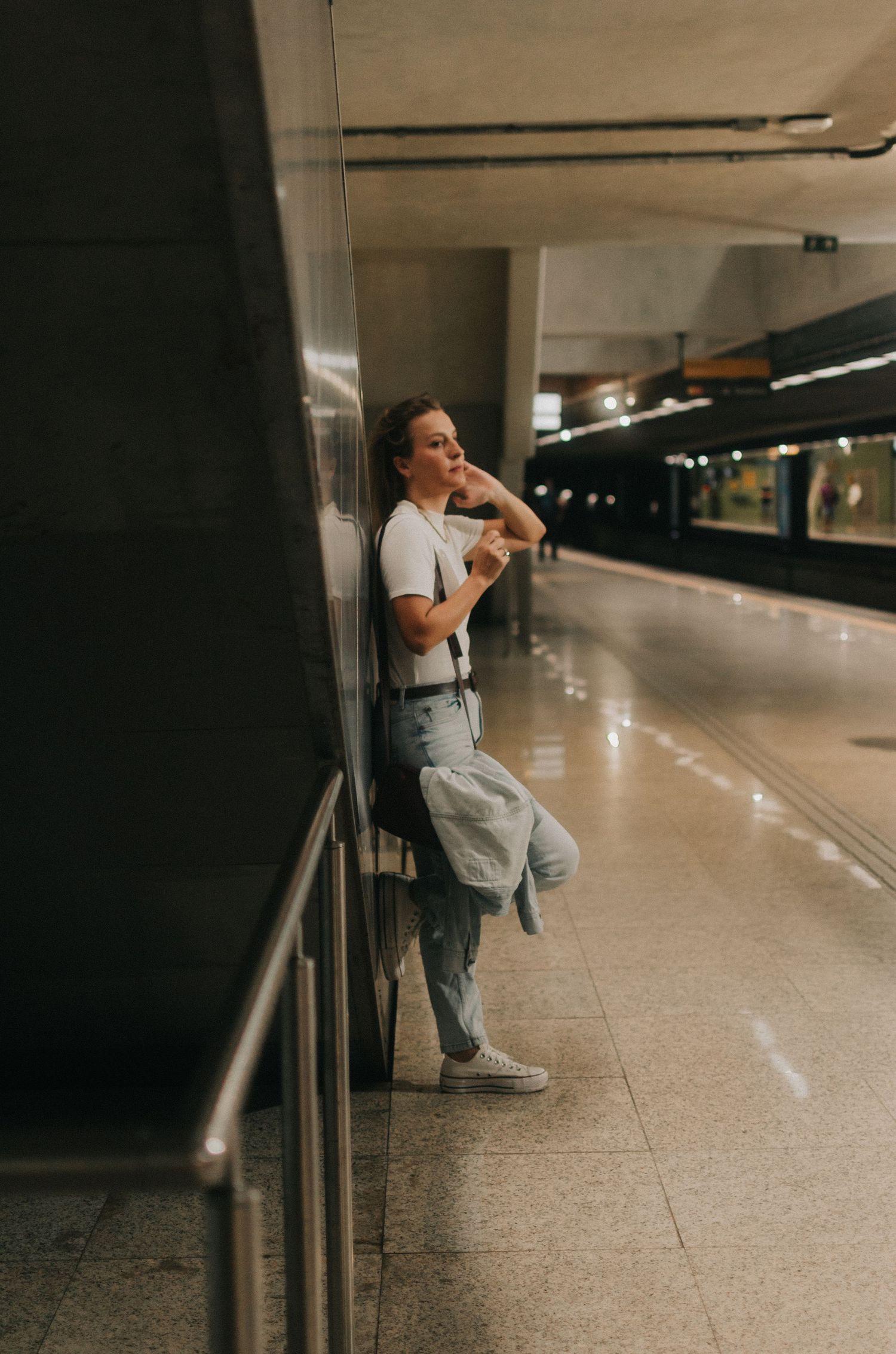 Person wearing jeans and white shirt leans against metal railing in empty subway station corridor.
