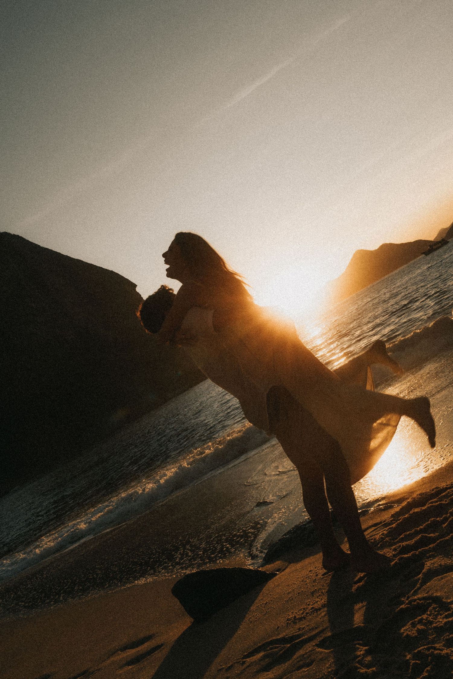 A silhouetted figure performs acrobatic moves on the beach during sunset with ocean waves in the background.