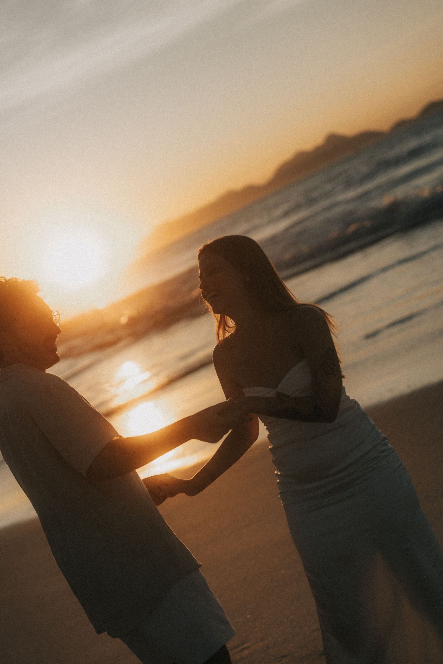 Two silhouettes dance together on a beach at sunset with ocean waves rolling in behind them in golden light.