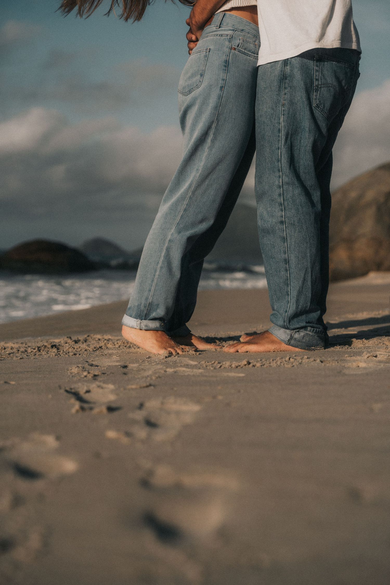 Multiple barefoot beach shots with someone in faded jeans walking along a sandy shore.