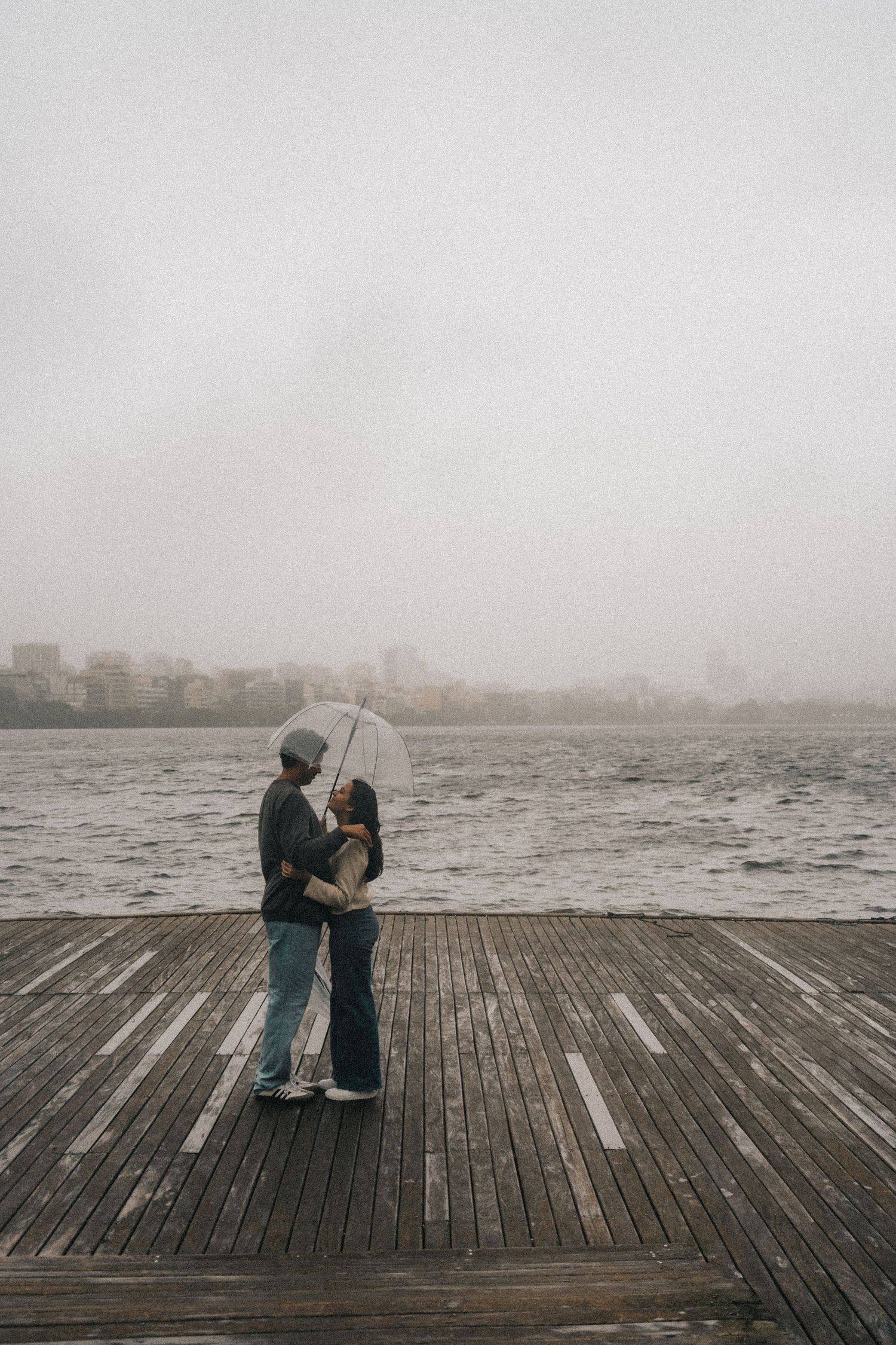 Two people stand together on a misty wooden boardwalk overlooking the sea in moody, dark weather.
