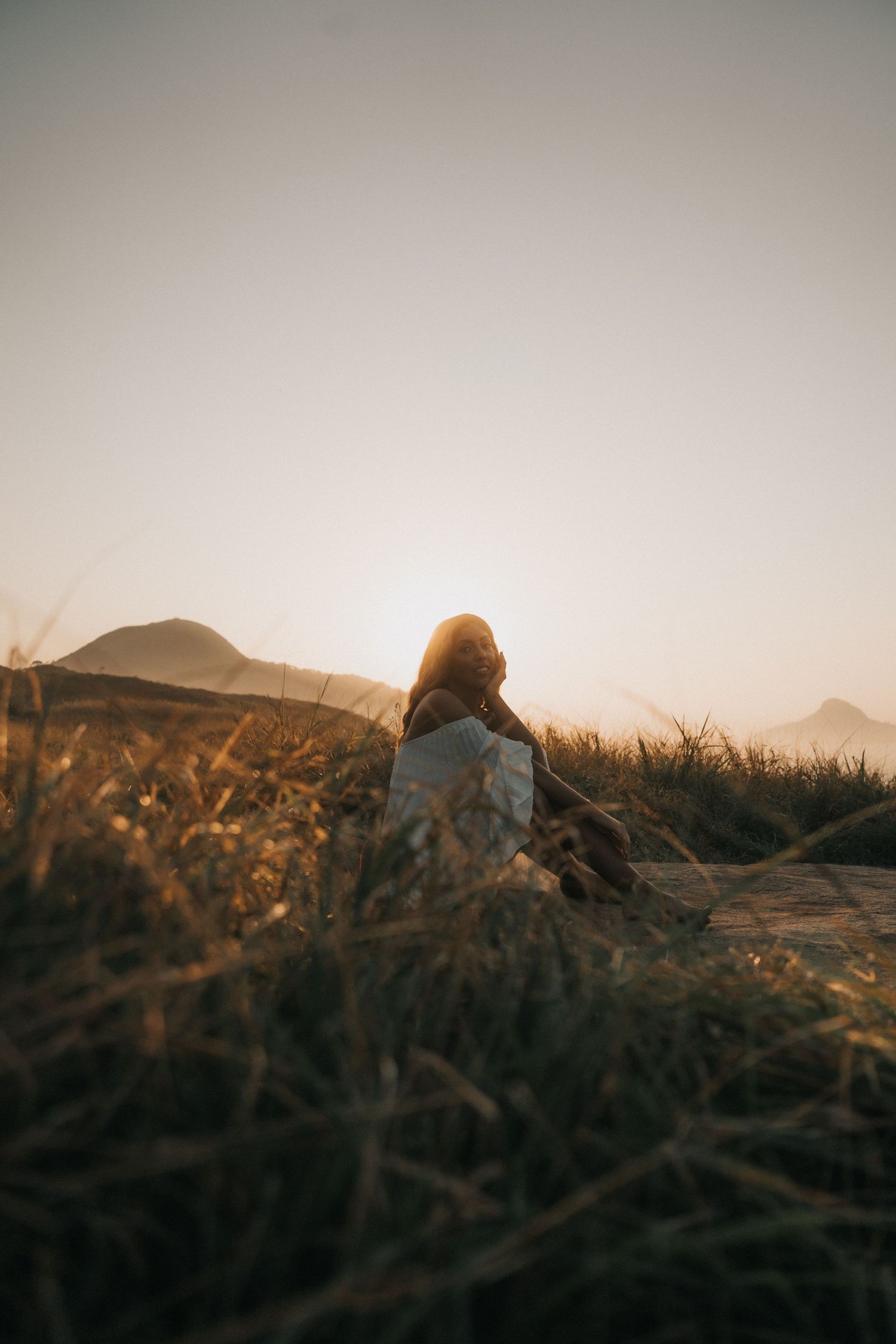 Silhouette of figure resting on grassy hillside at sunset with mountainous landscape in background and muted lighting.