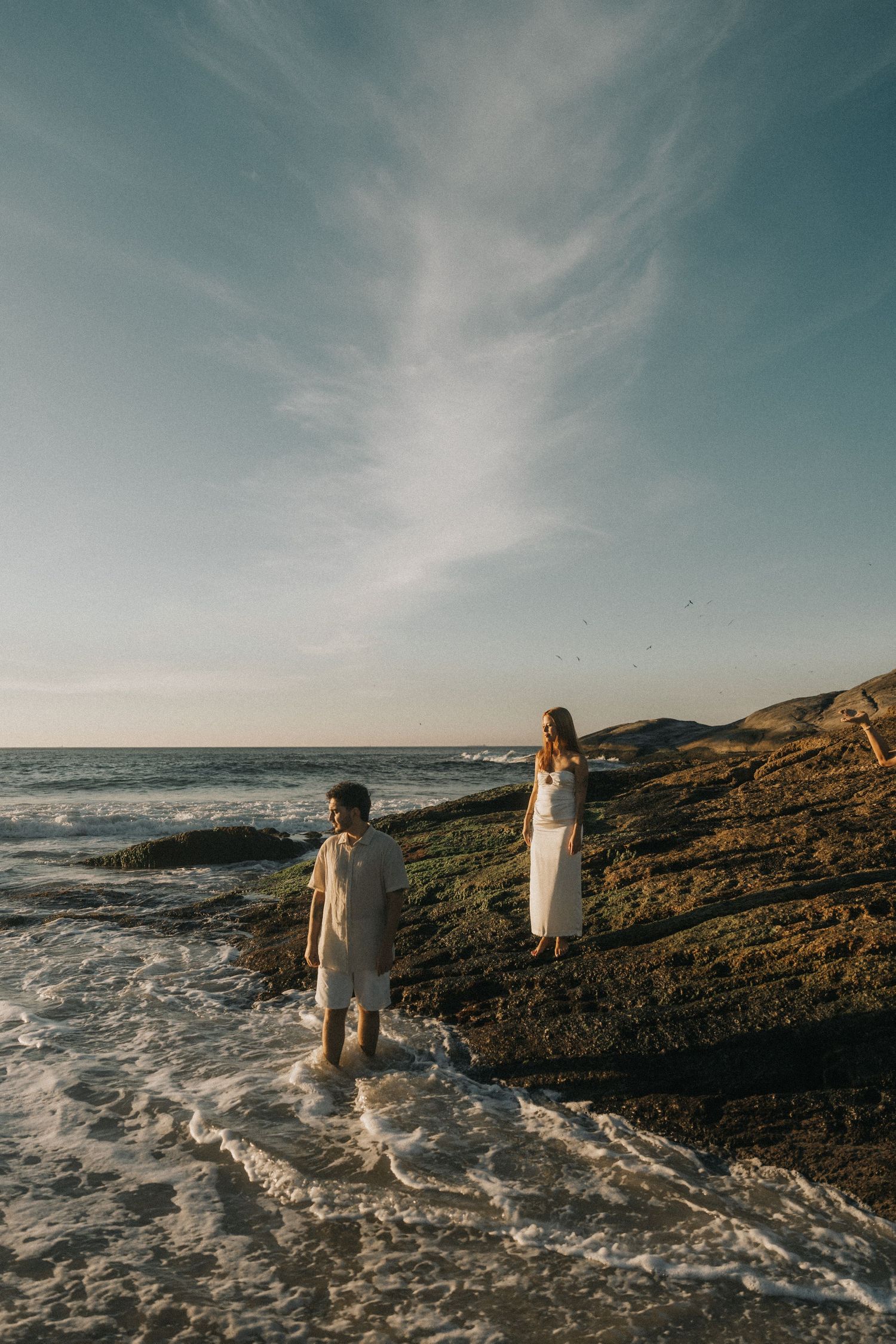 Two figures in light clothing stand on rugged coastal rocks as ocean waves crash against the shore during golden hour.