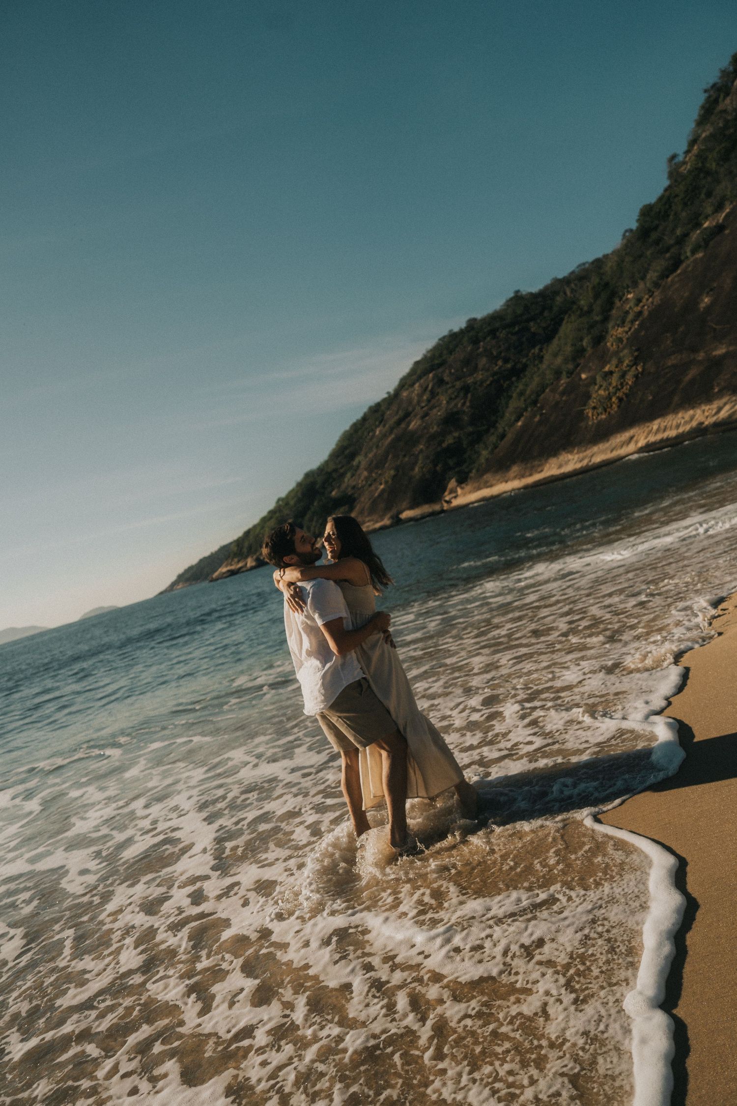 A couple embraces on a secluded beach with dramatic cliffs and crashing waves along the shoreline at sunset.