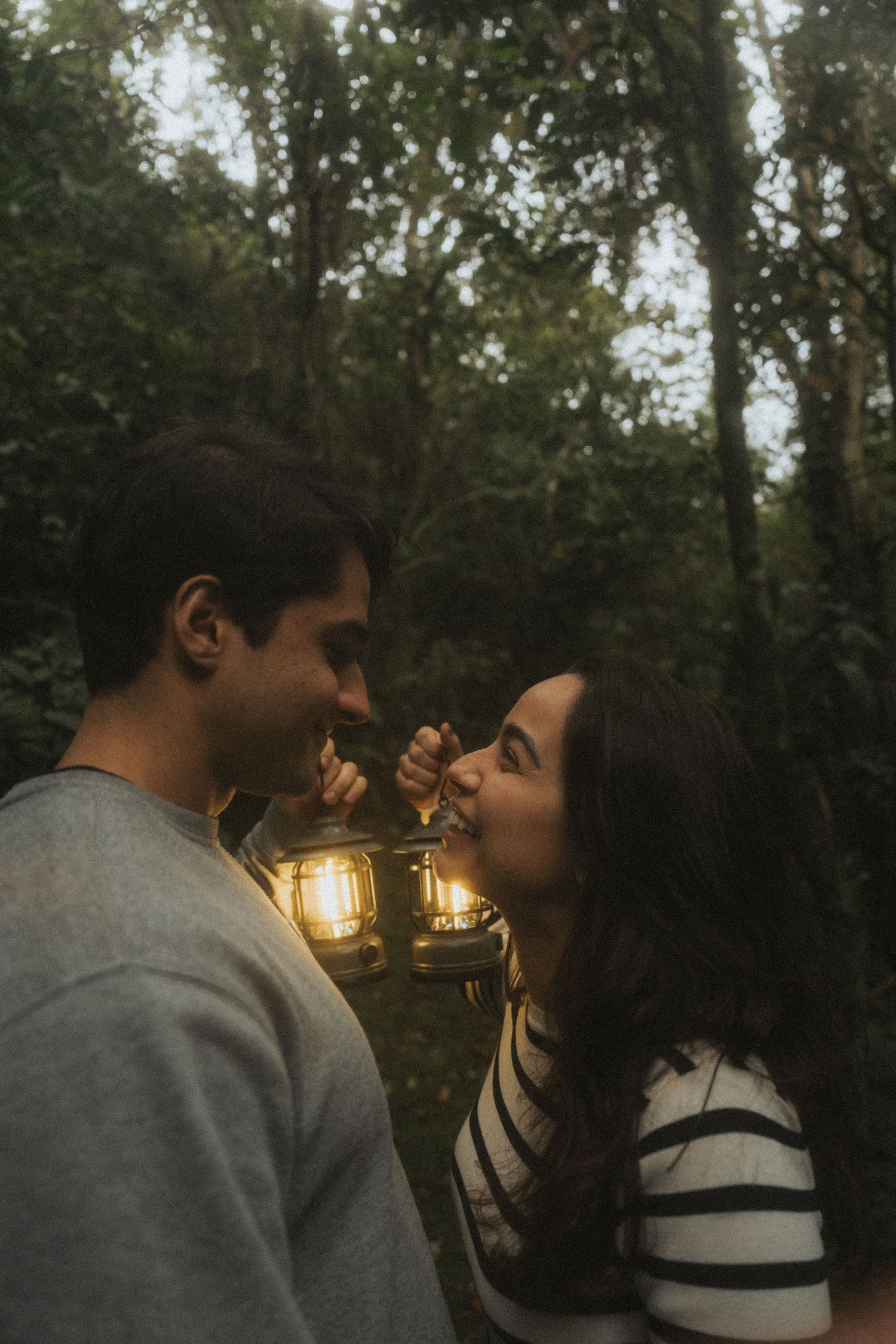 A romantic couple shares an intimate moment at sunset with glowing string lights in a wooded outdoor setting.