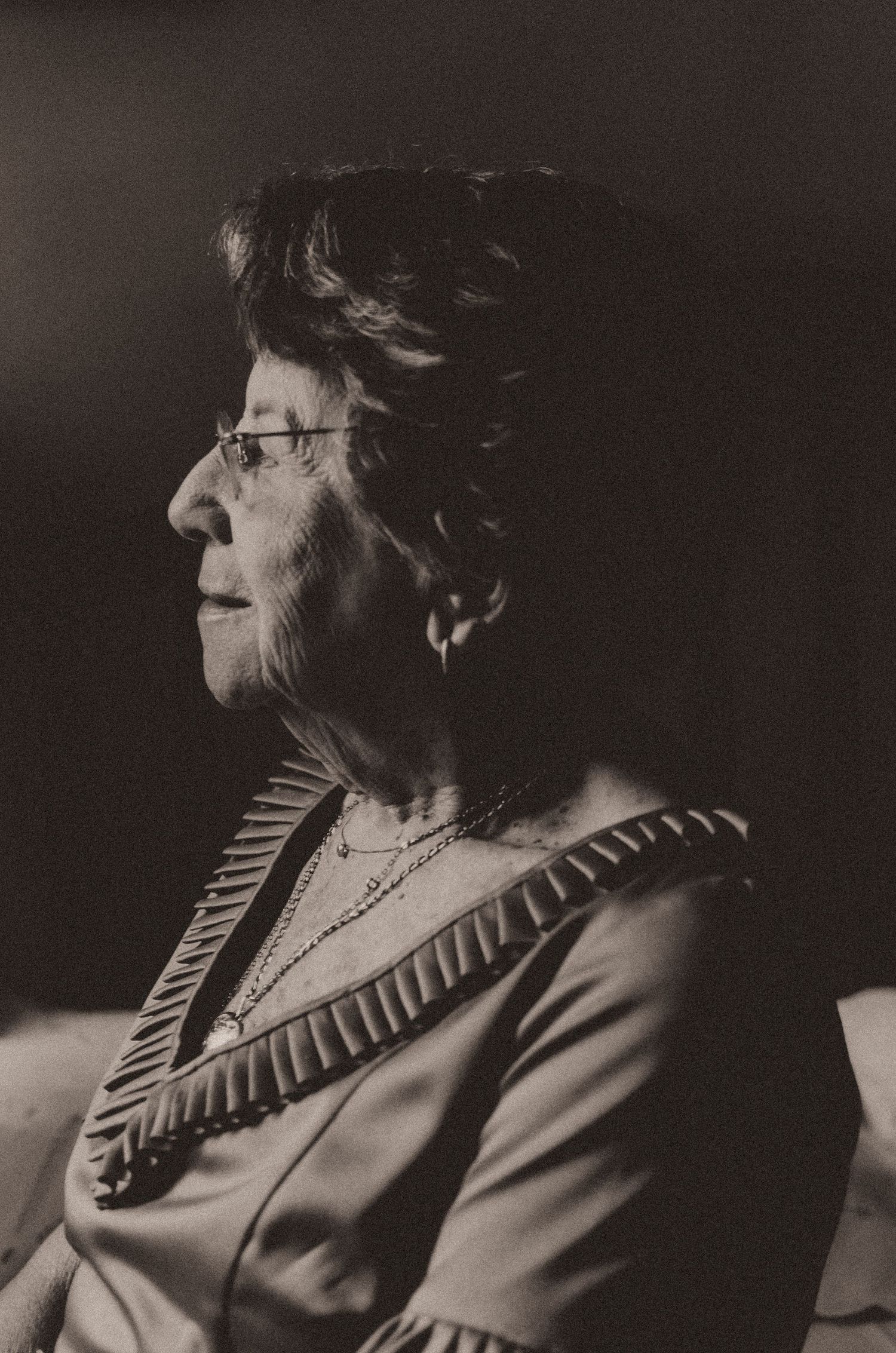 Black and white profile portrait of an elderly woman wearing glasses and a necklace against a dark background.