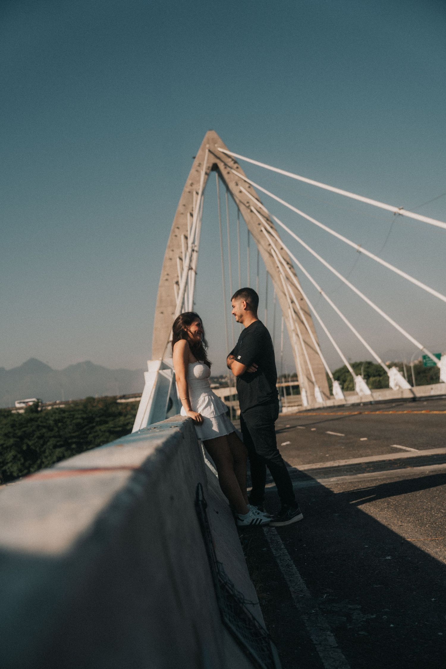 A couple stands together on a modern cable bridge at sunset with a dramatic teal sky in the background.