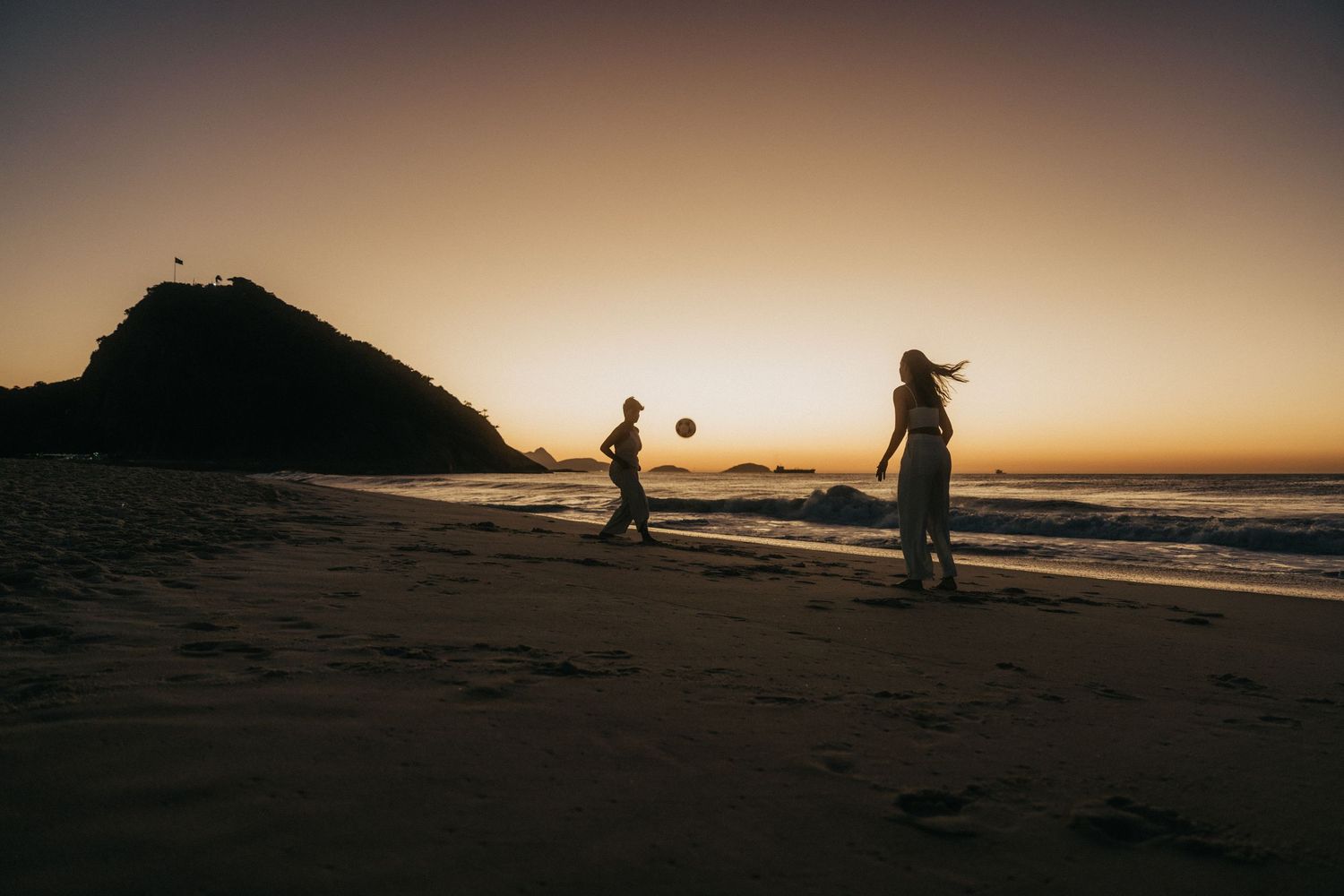 Silhouettes of people playing soccer on a beach at sunset against a dramatic orange and purple sky.